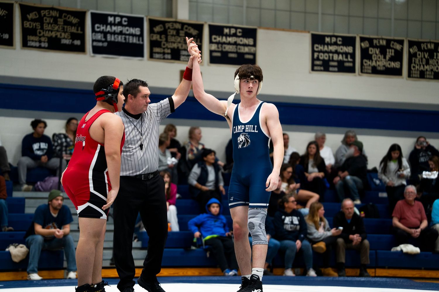 Referee raises hand of victorious Lions wrestler in blue singlet after winning match.