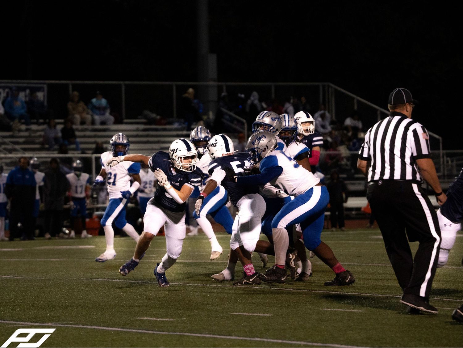 High school football teams in navy and blue uniforms battle at line of scrimmage during night game.