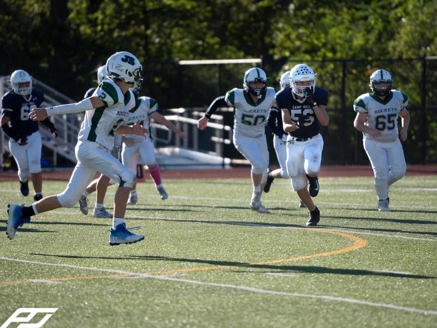 Green-uniformed football player runs with ball as navy-uniformed defenders give chase on sunny turf field.