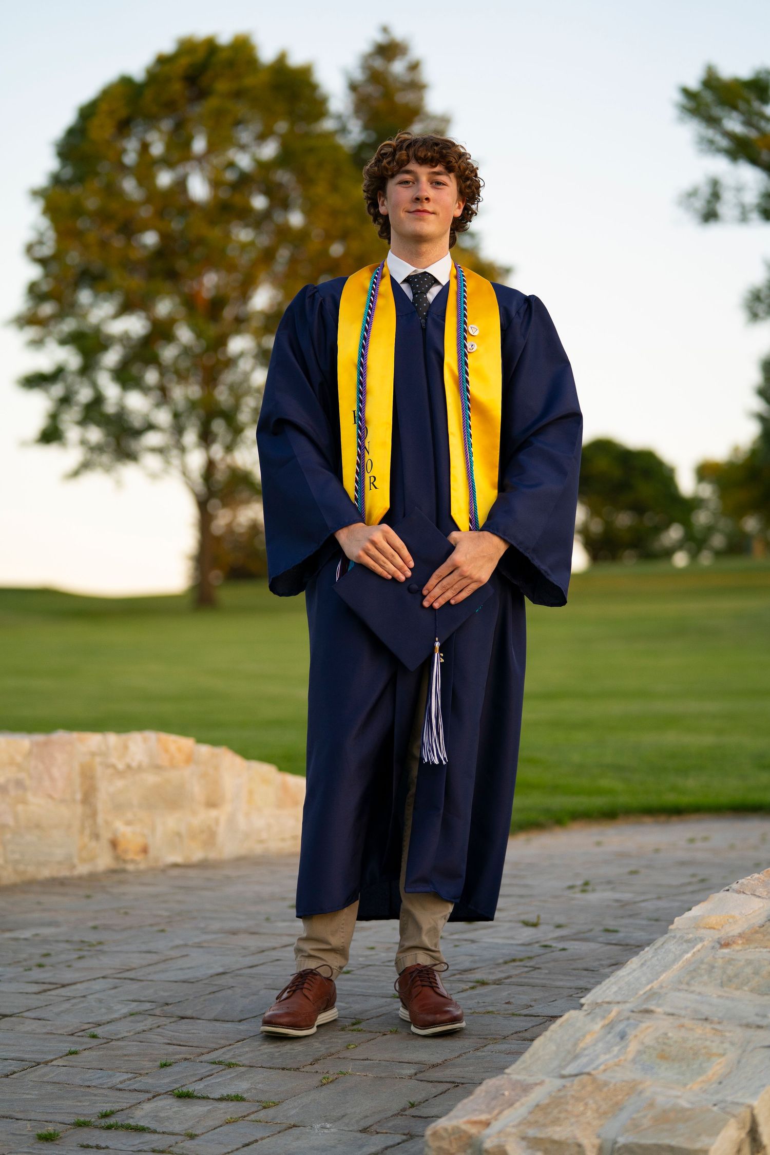 Young male graduate in navy and gold graduation gown stands confidently on stone pathway outdoors.