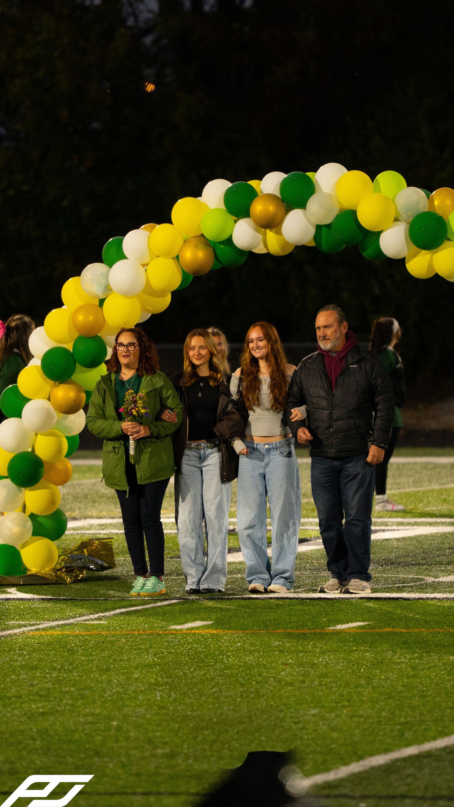Four people walk under a green, yellow, and white balloon arch during a high school senior night celebration.