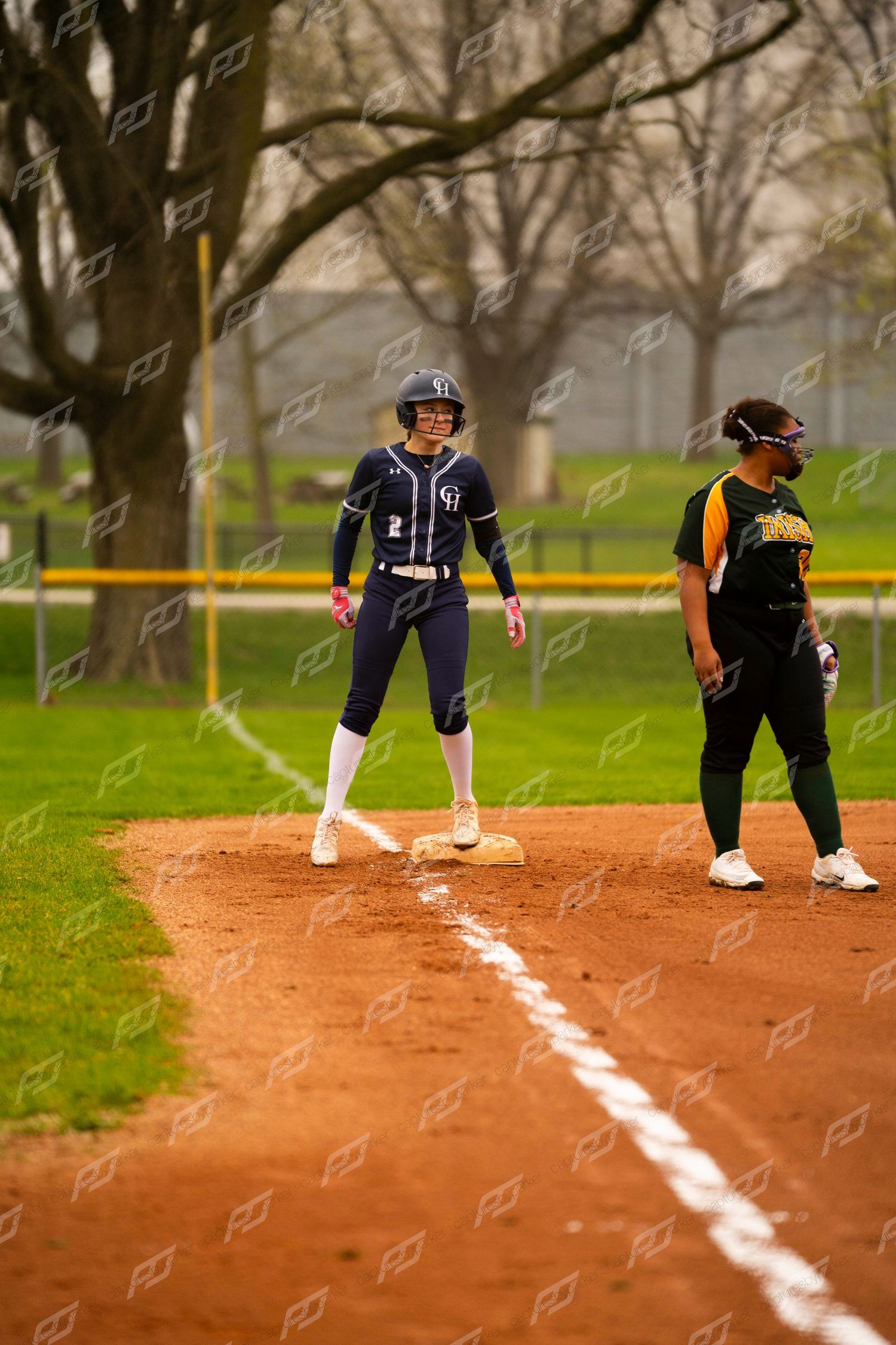 Softball player number 2 in navy uniform stands on base next to opposing team fielder on clay infield.