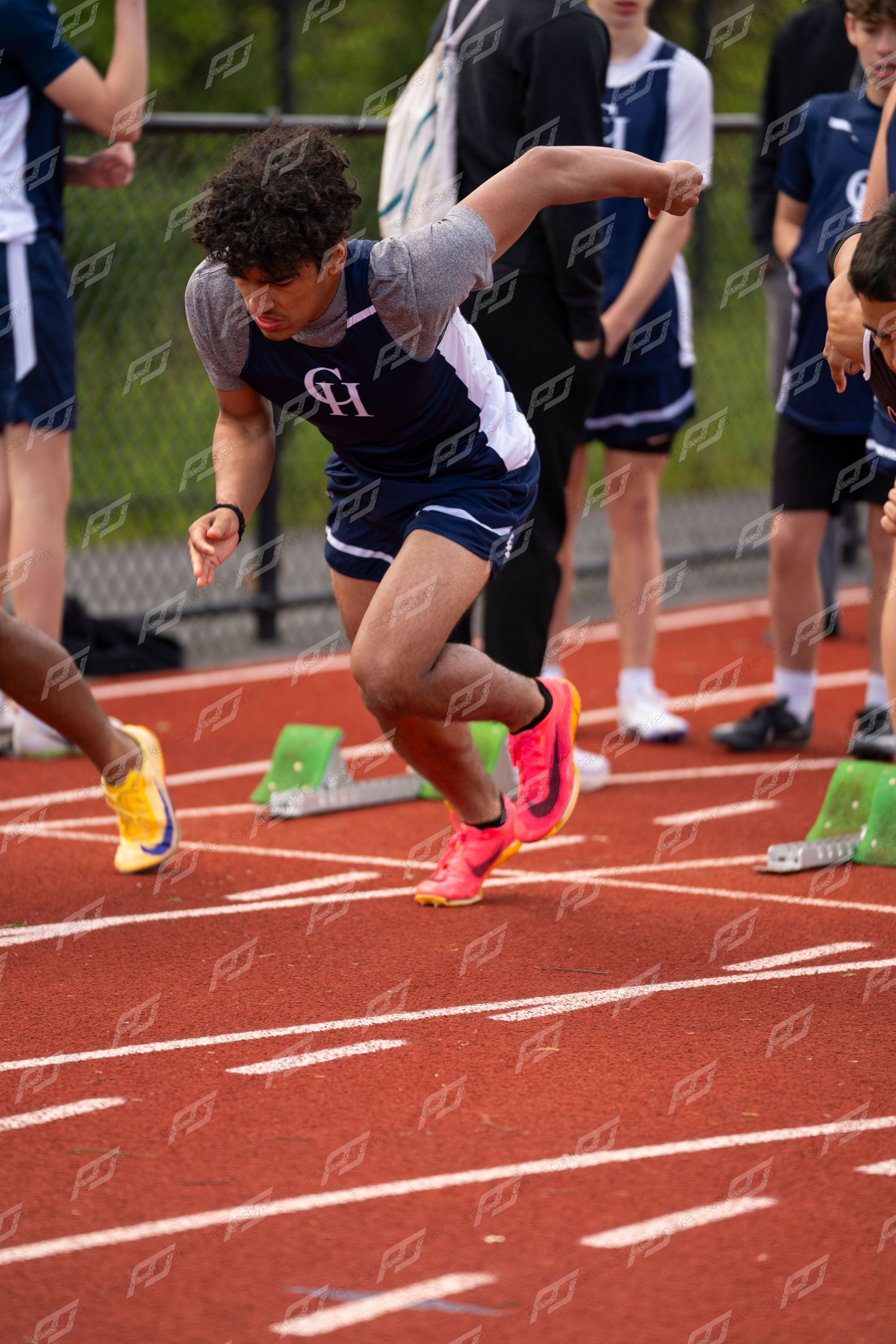 Male sprinter explodes from starting blocks in navy uniform at outdoor track competition.