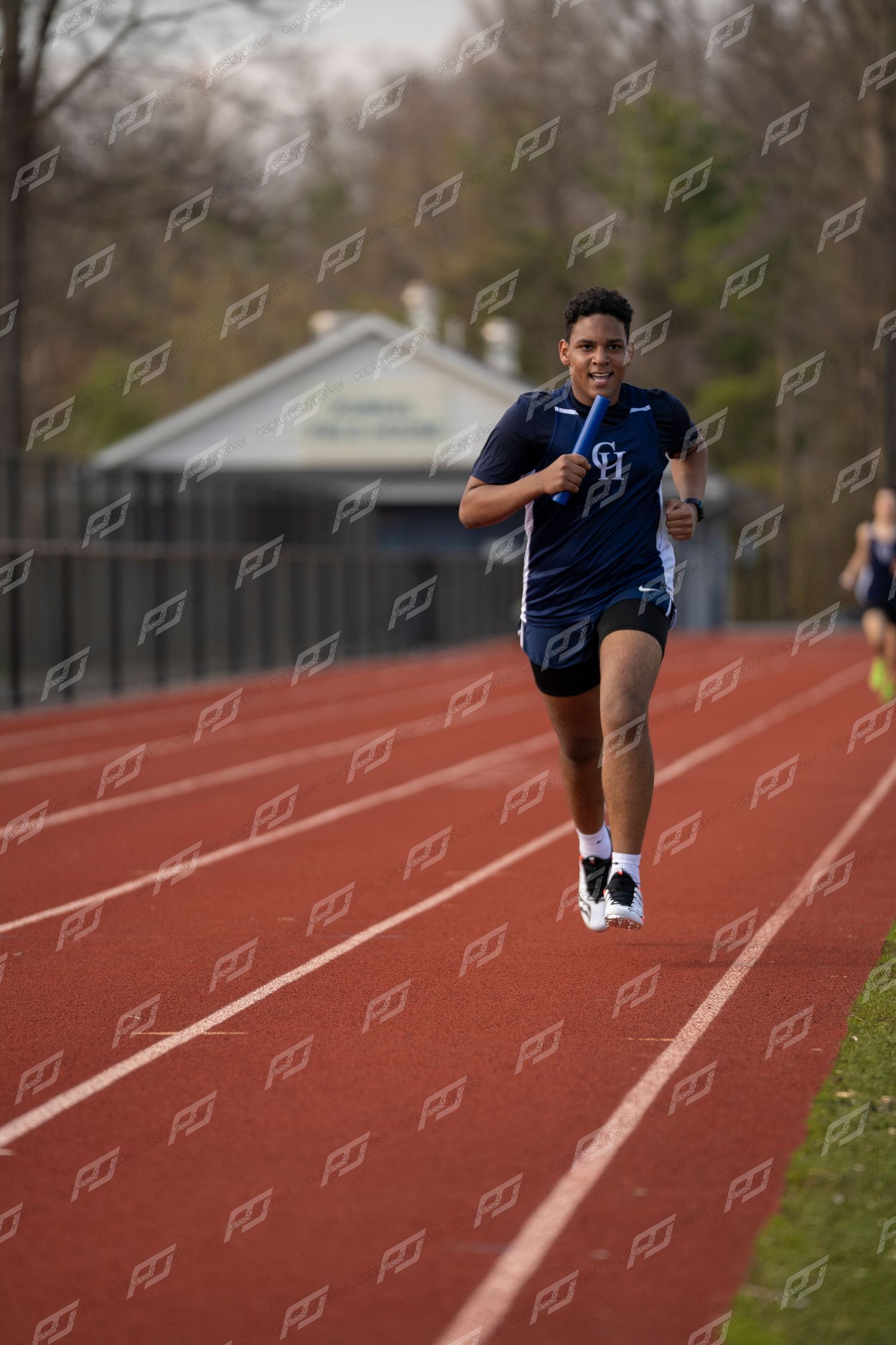 Smiling male high school runner in navy CH jersey sprints on red outdoor track.