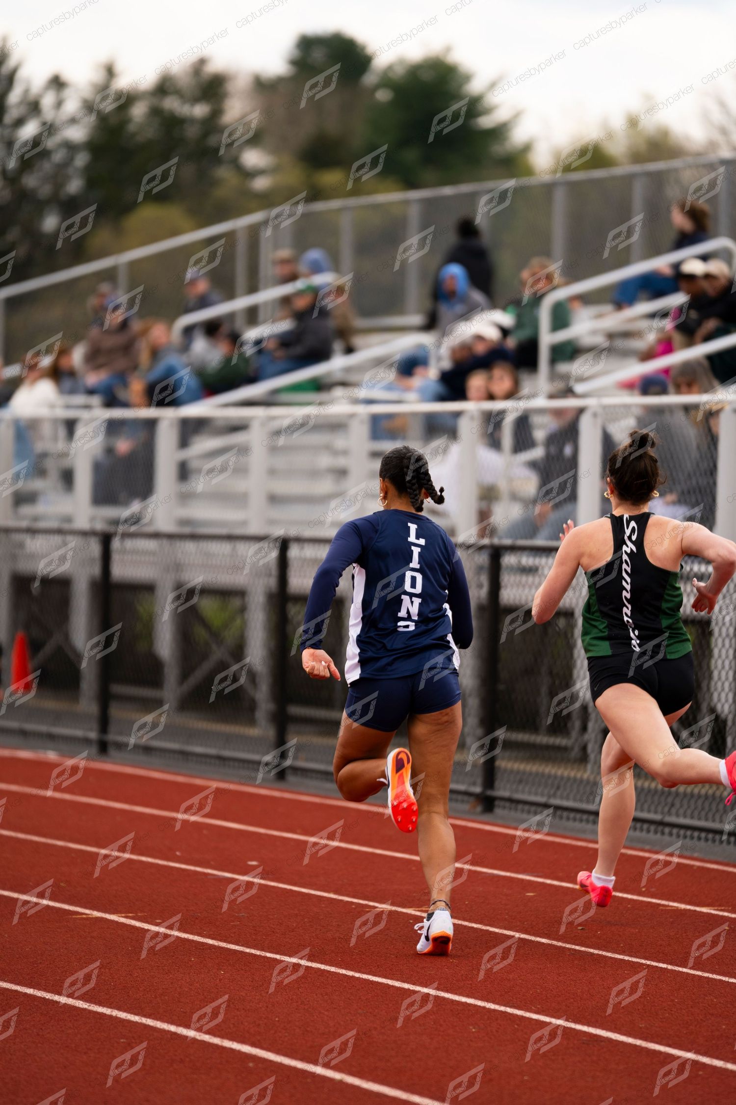 Two female sprinters race on red track during outdoor high school track and field competition.