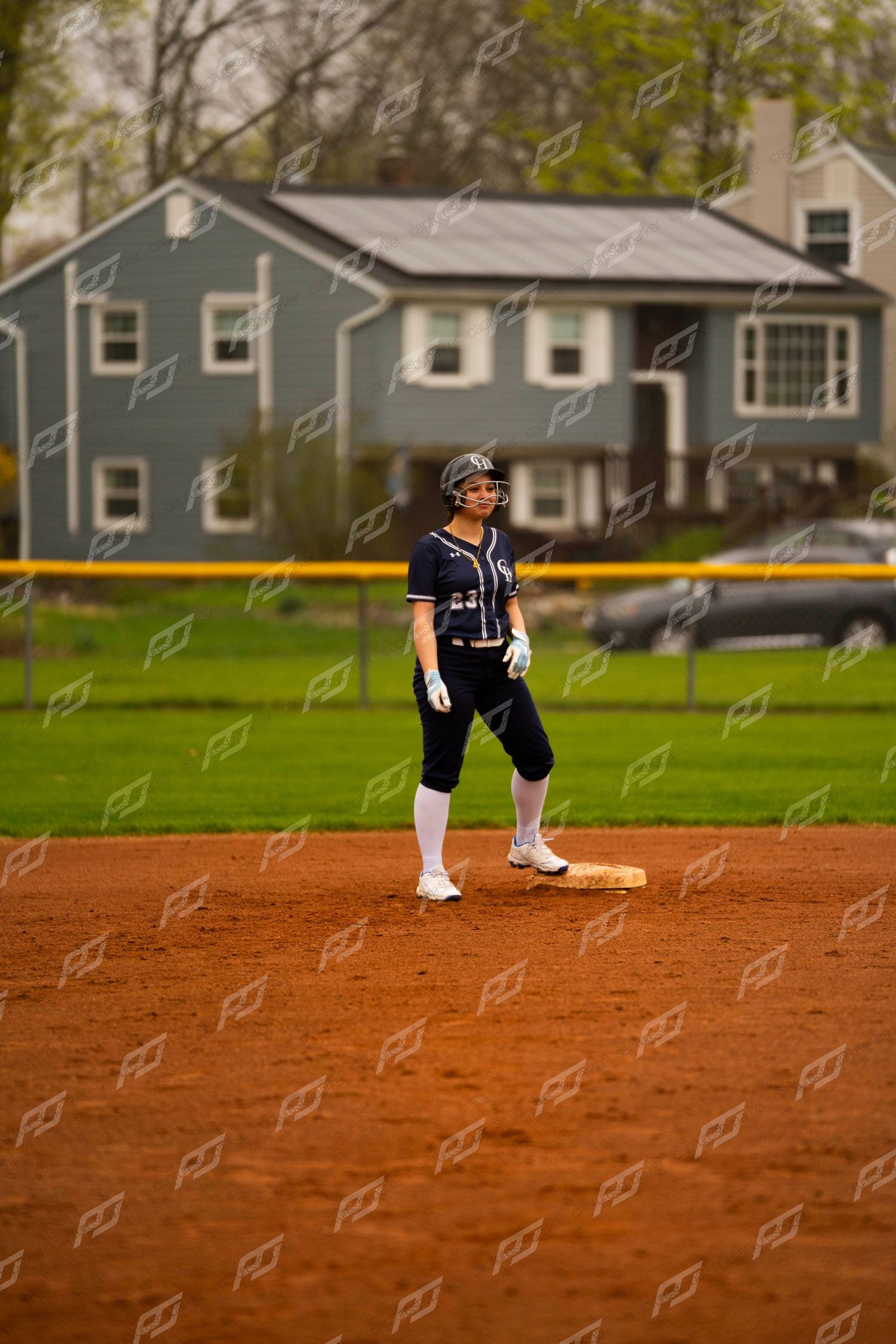 Female softball player in navy uniform stands alert on base during outdoor game on dirt field.