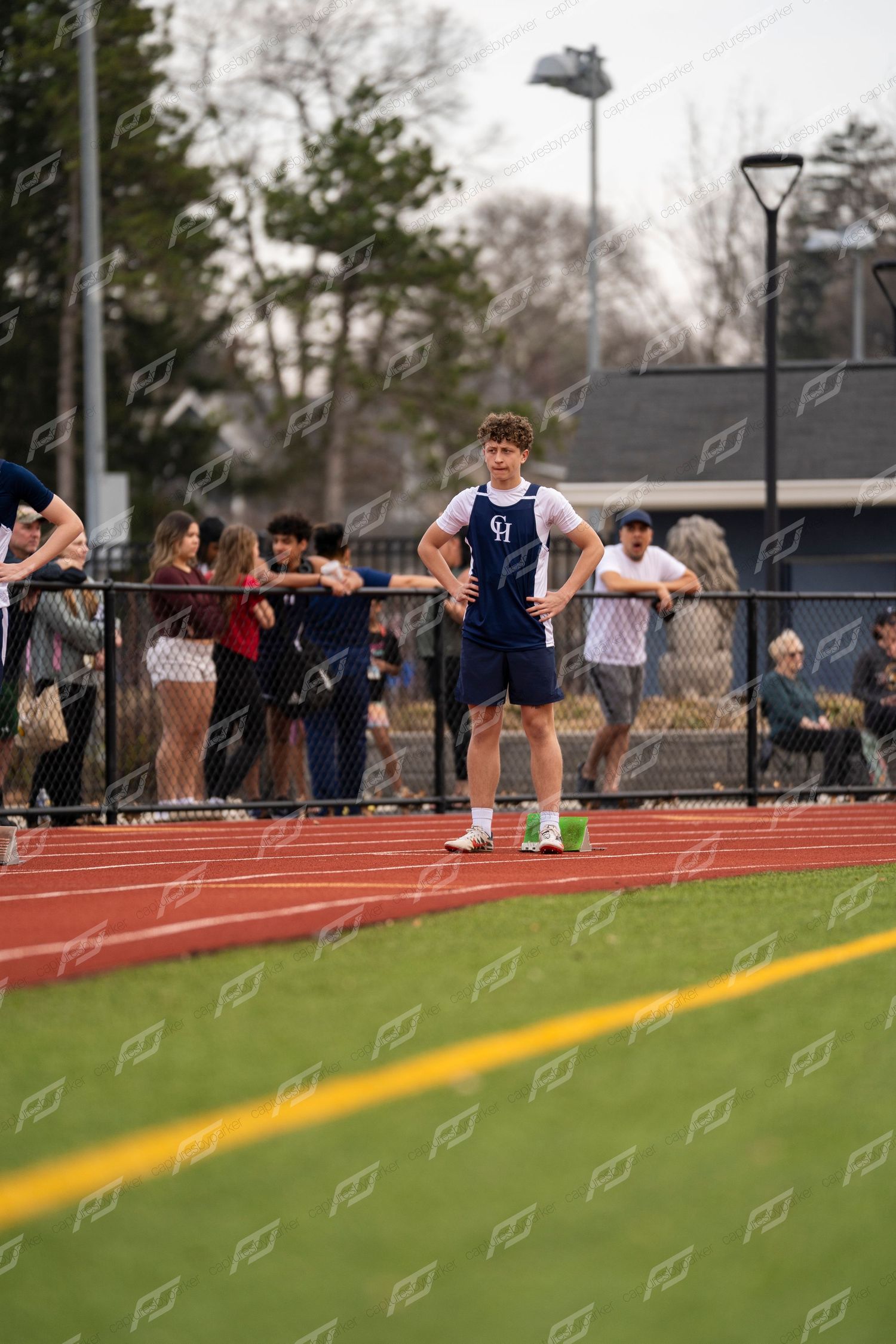 Male track athlete in navy uniform stands with hands on hips at track meet.