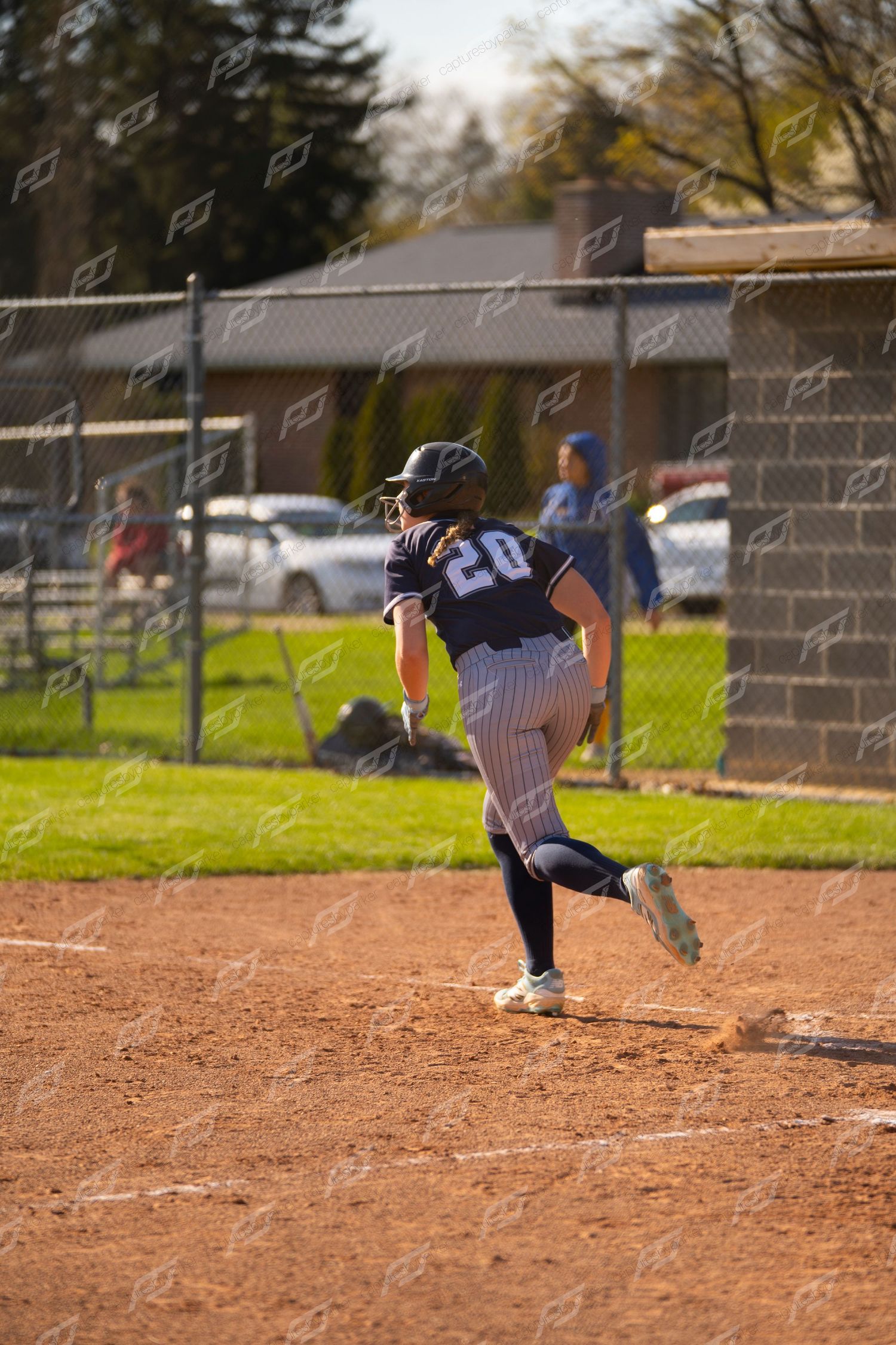 Female softball player #20 in navy pinstripe uniform running bases on dirt infield during game.
