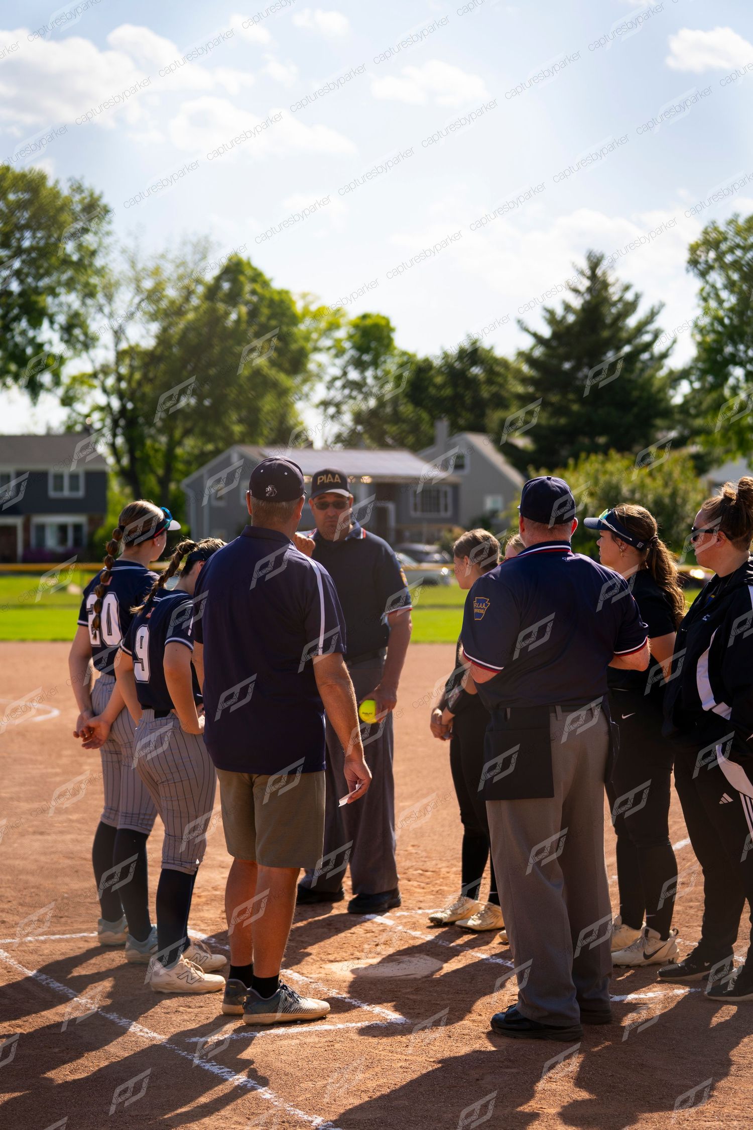 Softball team huddles with coaches at home plate during a sunny game day.