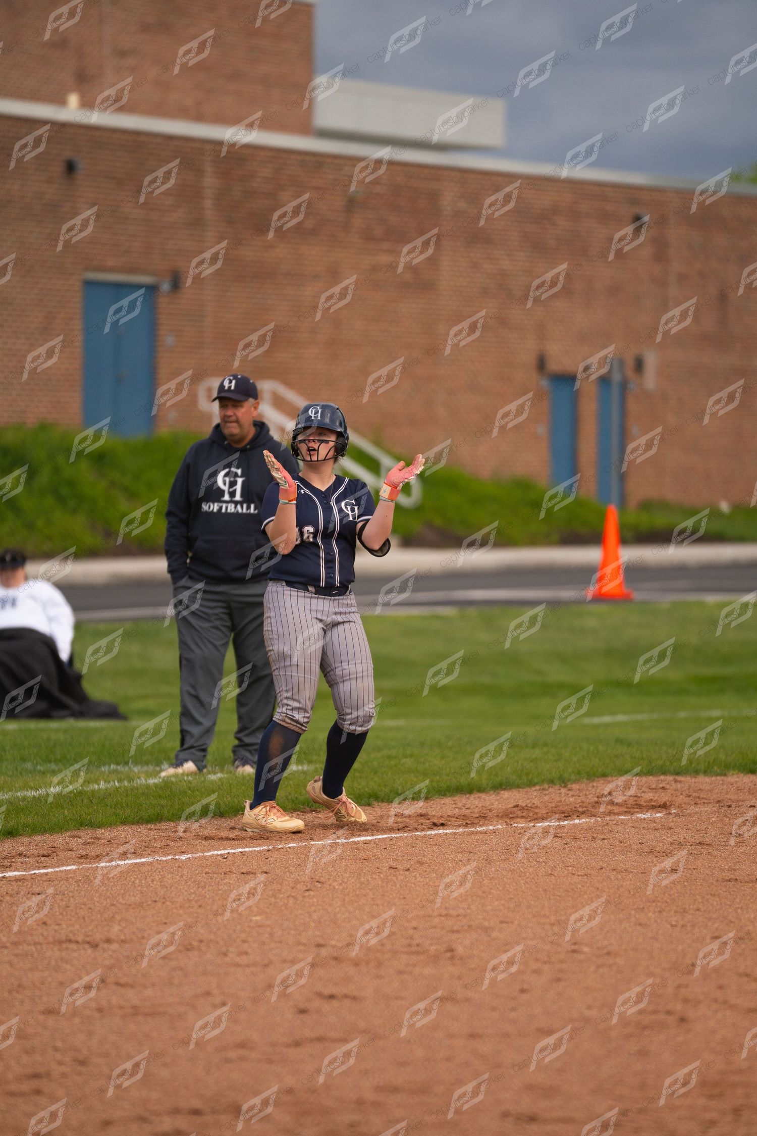 Softball player celebrates on base with a coach wearing a GH Softball jacket nearby.