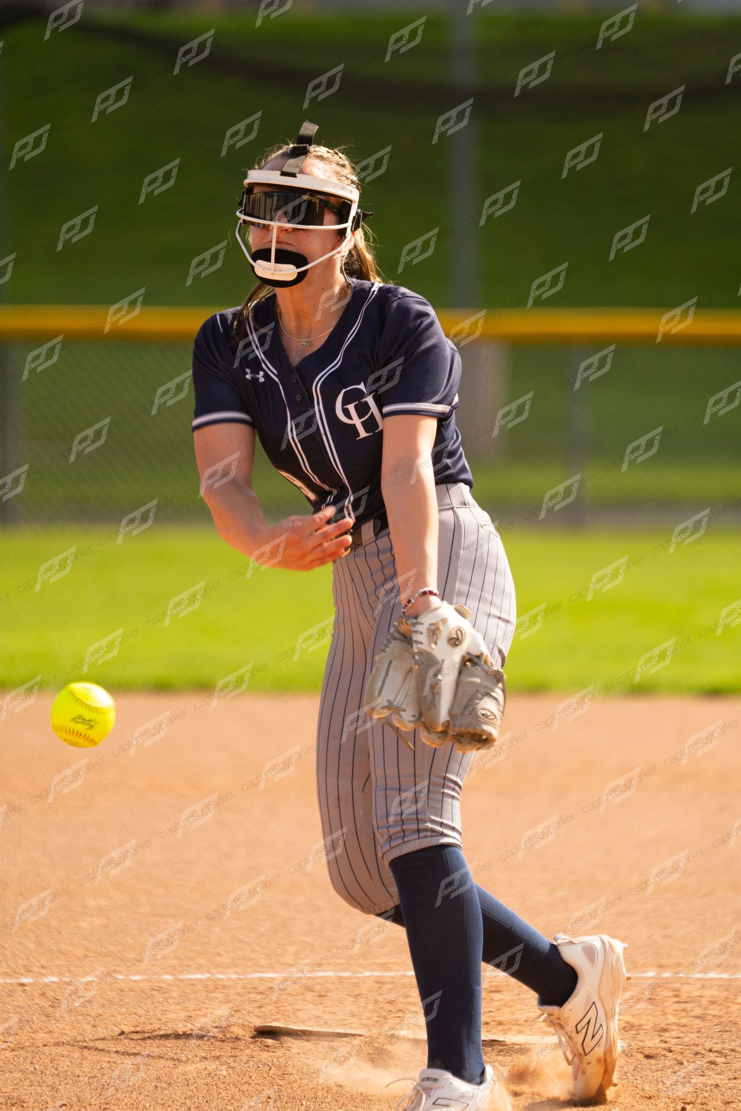 Female softball pitcher in navy pinstripe uniform delivers a pitch on a sunny day.