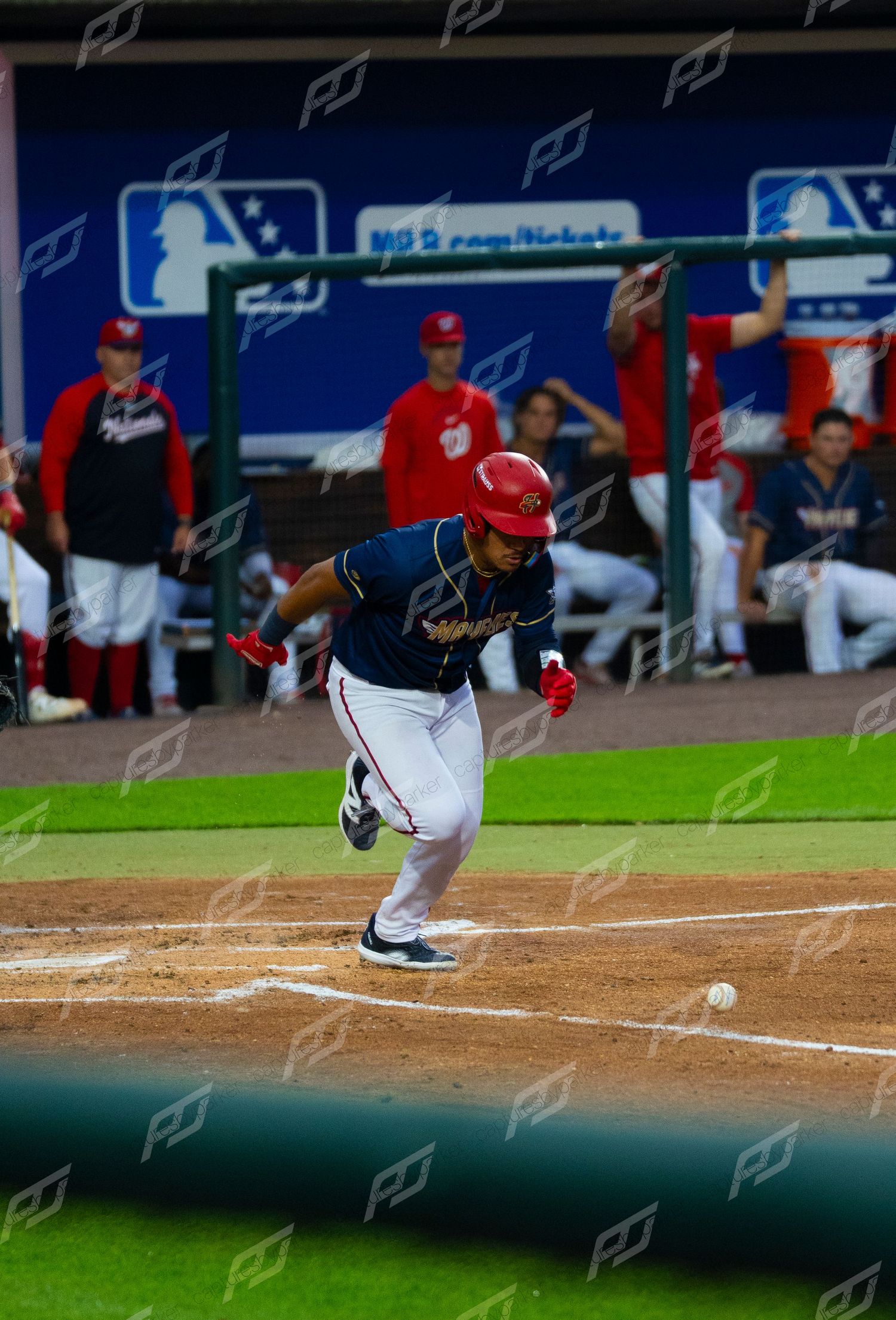 Minor league baseball player sprints from home plate after making contact as teammates cheer from dugout.