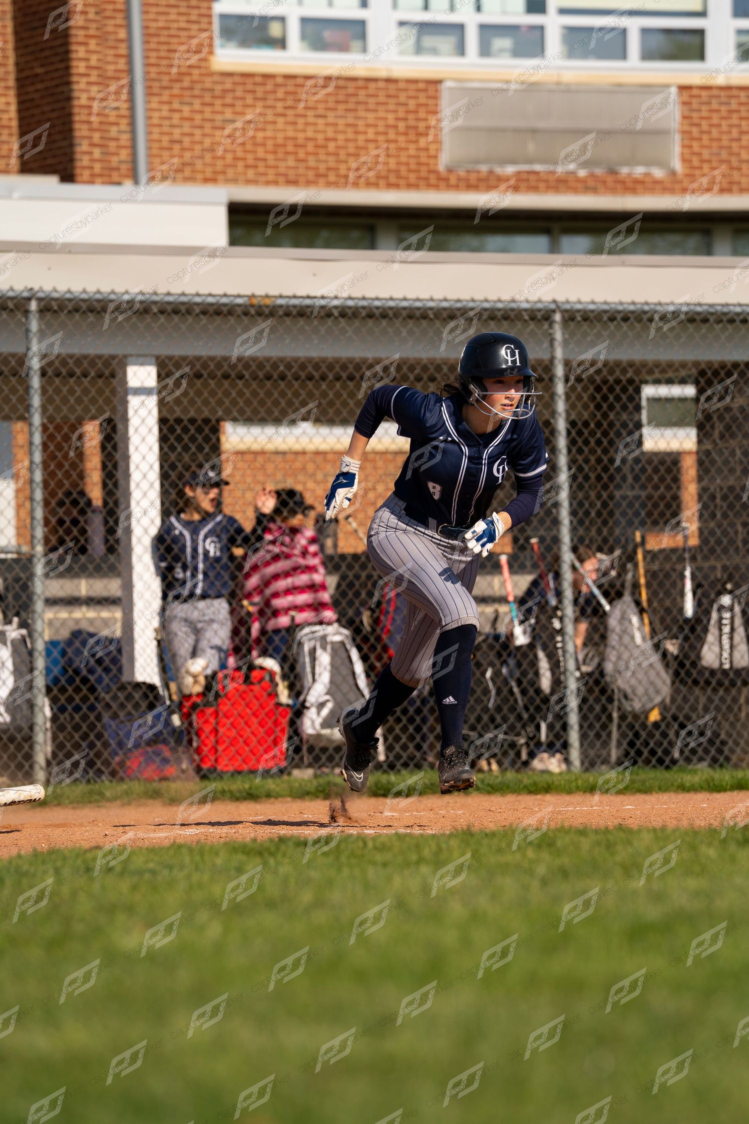 Softball player in navy uniform sprinting away from home plate after making contact during a game.