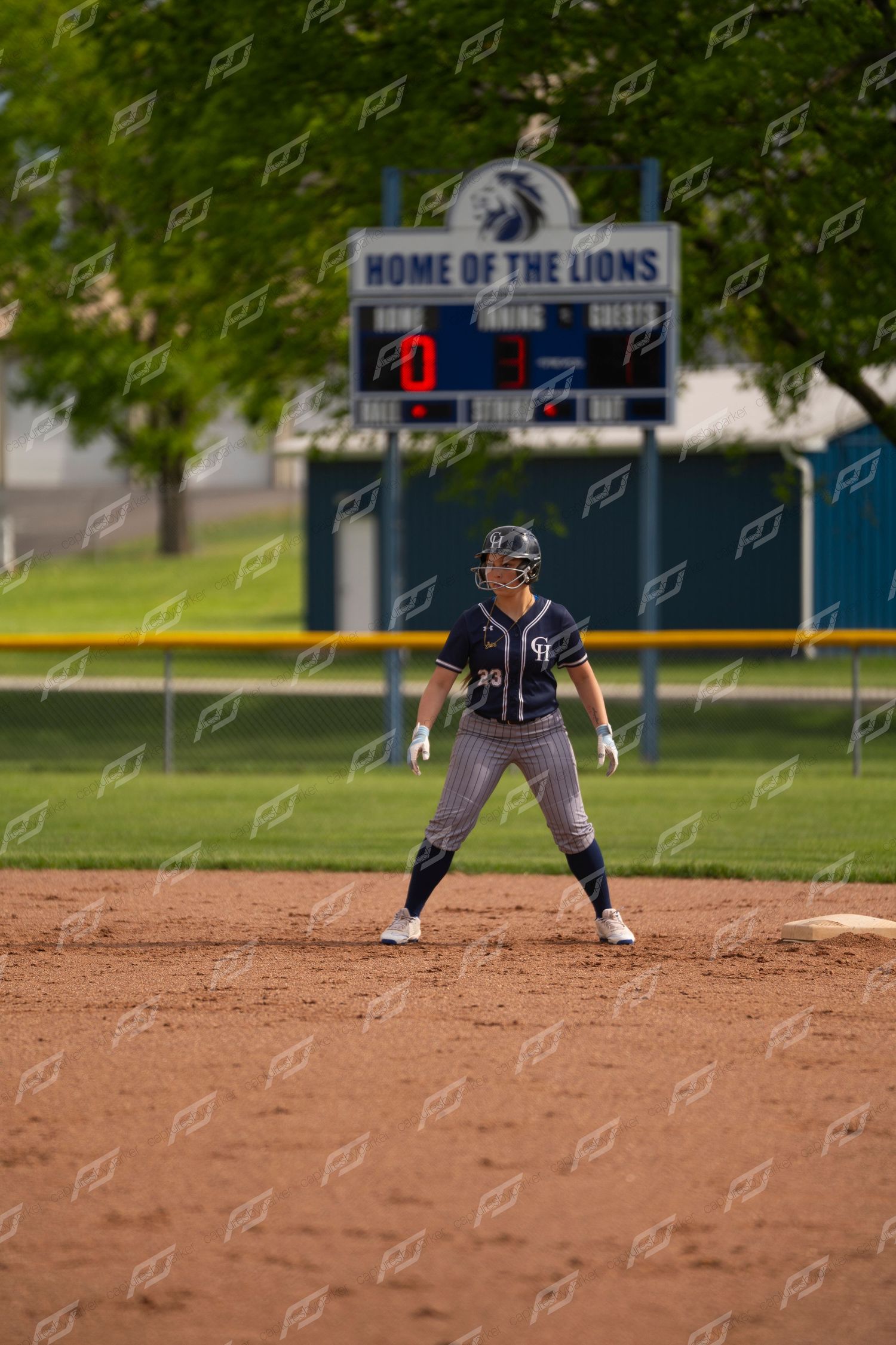 Softball player in navy uniform leads off base at Home of the Lions field with scoreboard visible.