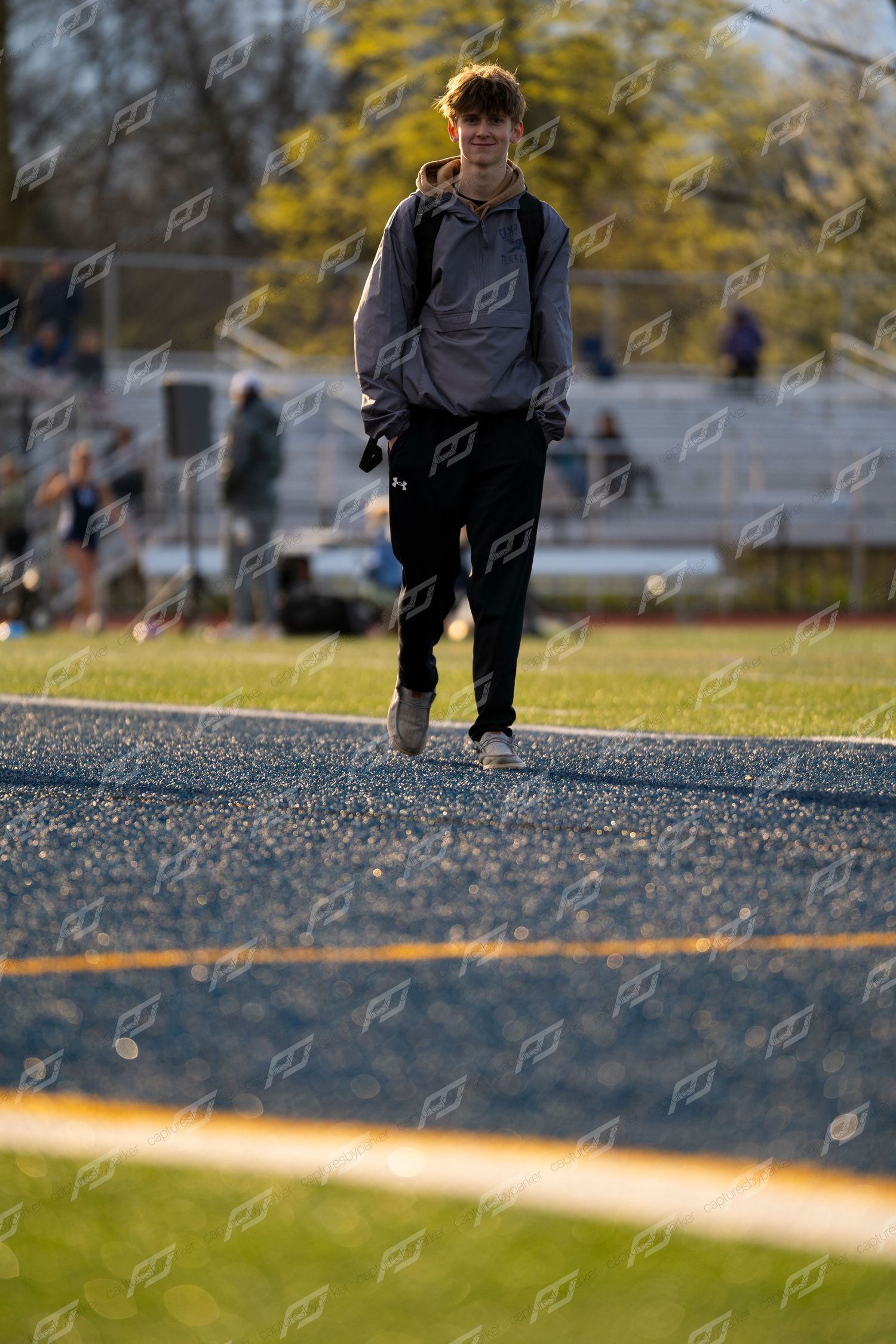 Young male athlete in gray hoodie walks along blue track at outdoor sports facility.