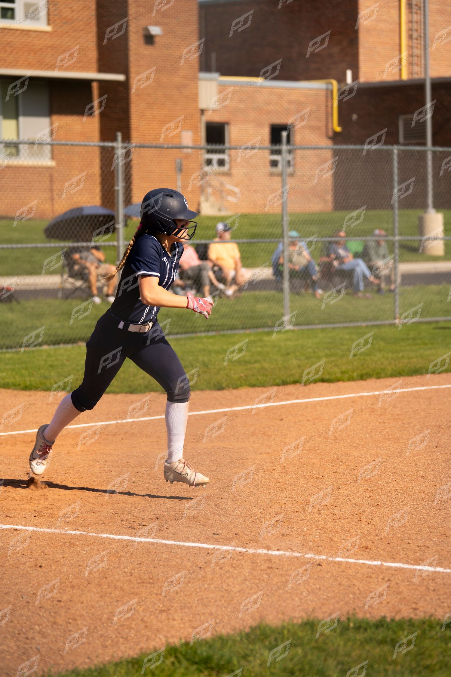 Softball player in navy uniform running the bases on a sunny day during a game.