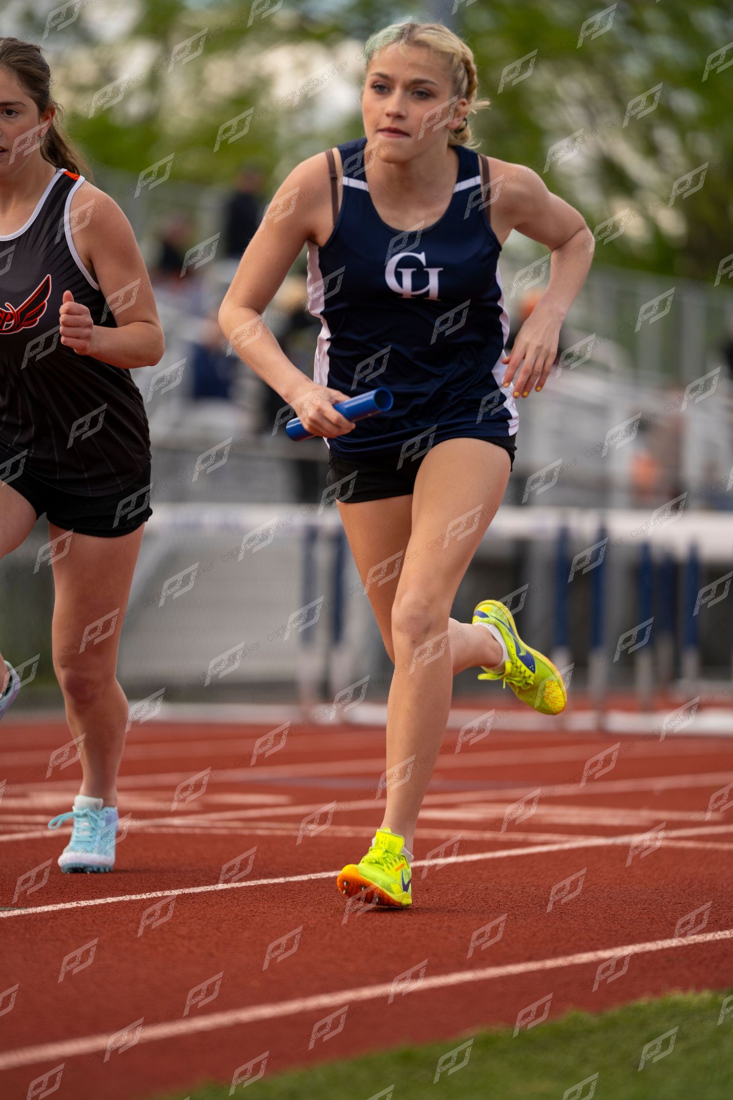 Female track athlete in a GH uniform running a relay race, holding a blue baton on a red outdoor track.