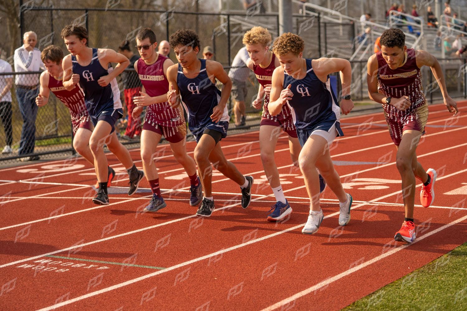 Group of male track athletes in navy CH uniforms starting a race on red track.