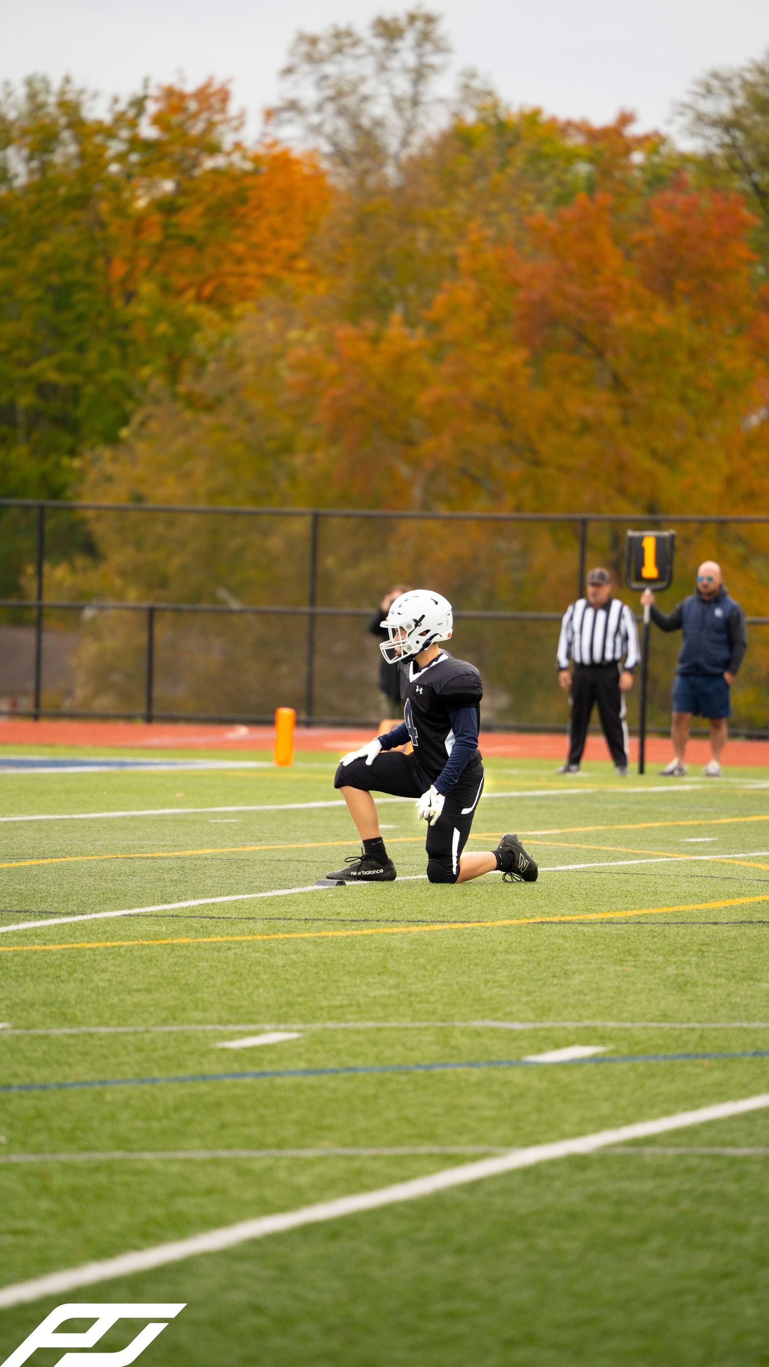 Young football player in black uniform kneels on green turf field during an autumn youth football game.