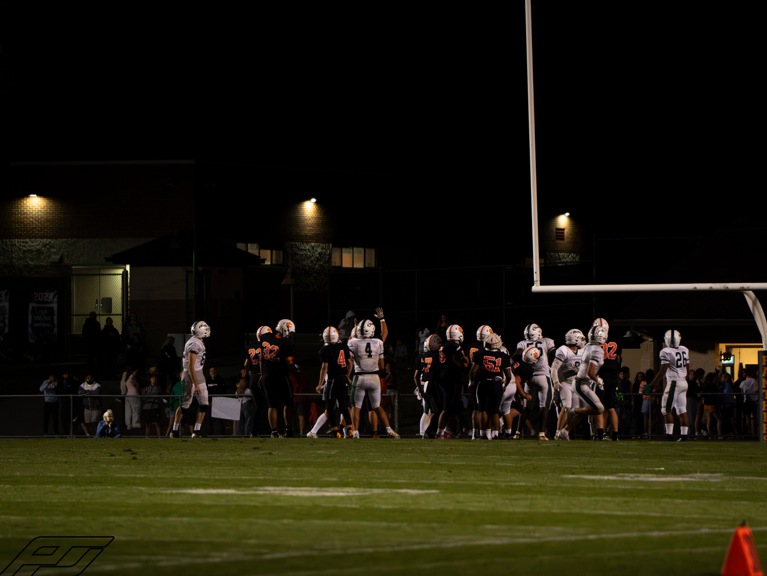High school football teams gather near the goal post under bright stadium lights during a night game.