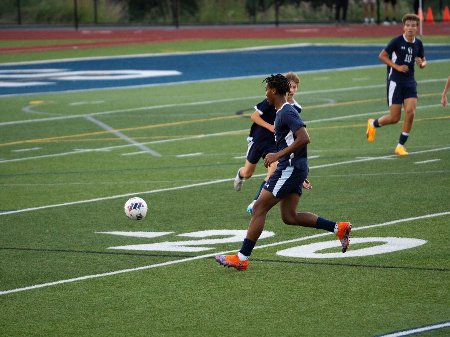 Soccer player in navy uniform dribbles ball down green turf field during evening match.