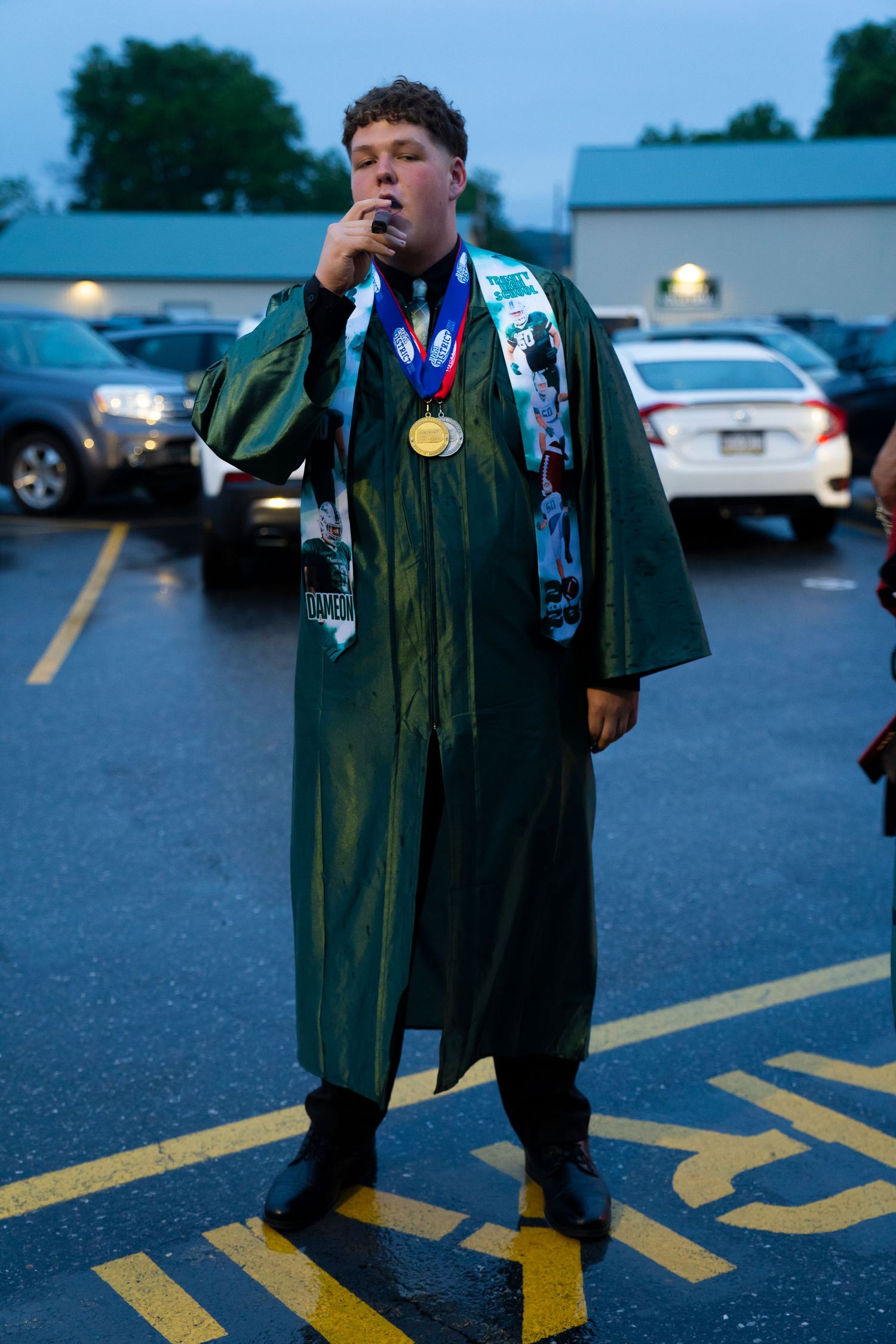 Graduate in green cap and gown wearing gold medal poses in parking lot at dusk.