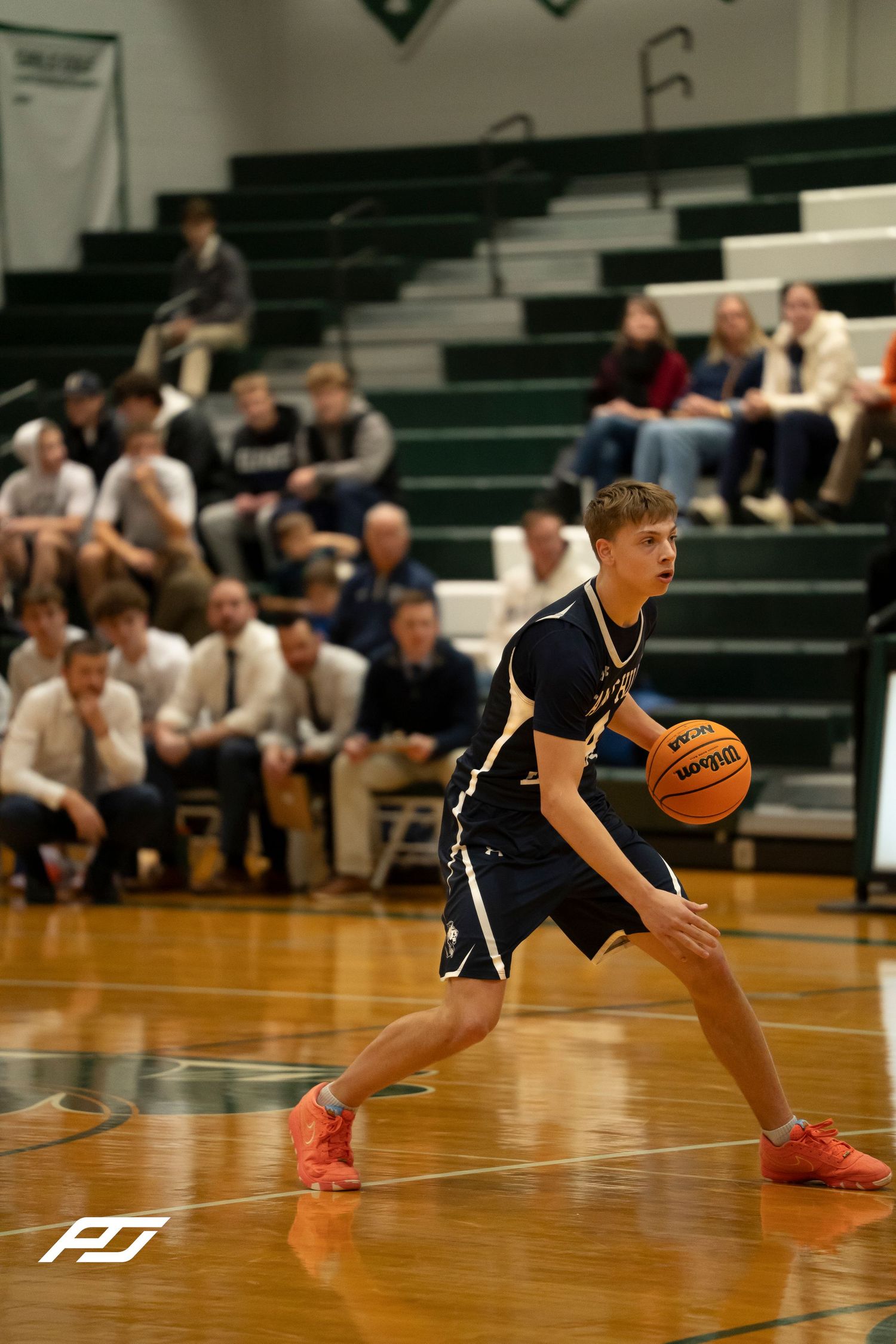 Teen basketball player dribbles ball in navy uniform during high school game with crowded bleachers.