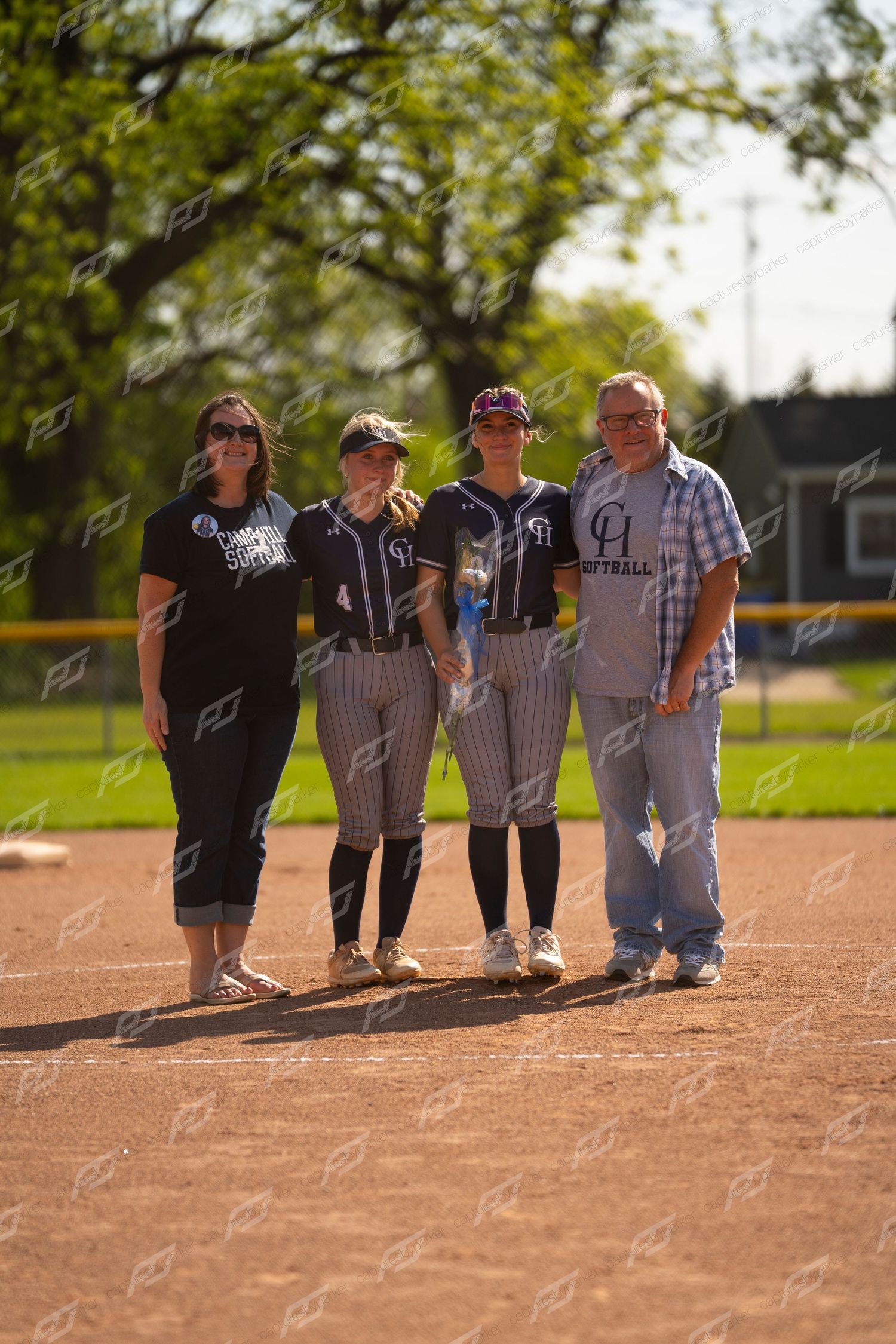 Two softball players in navy uniforms stand with parents on baseball field during senior day ceremony.
