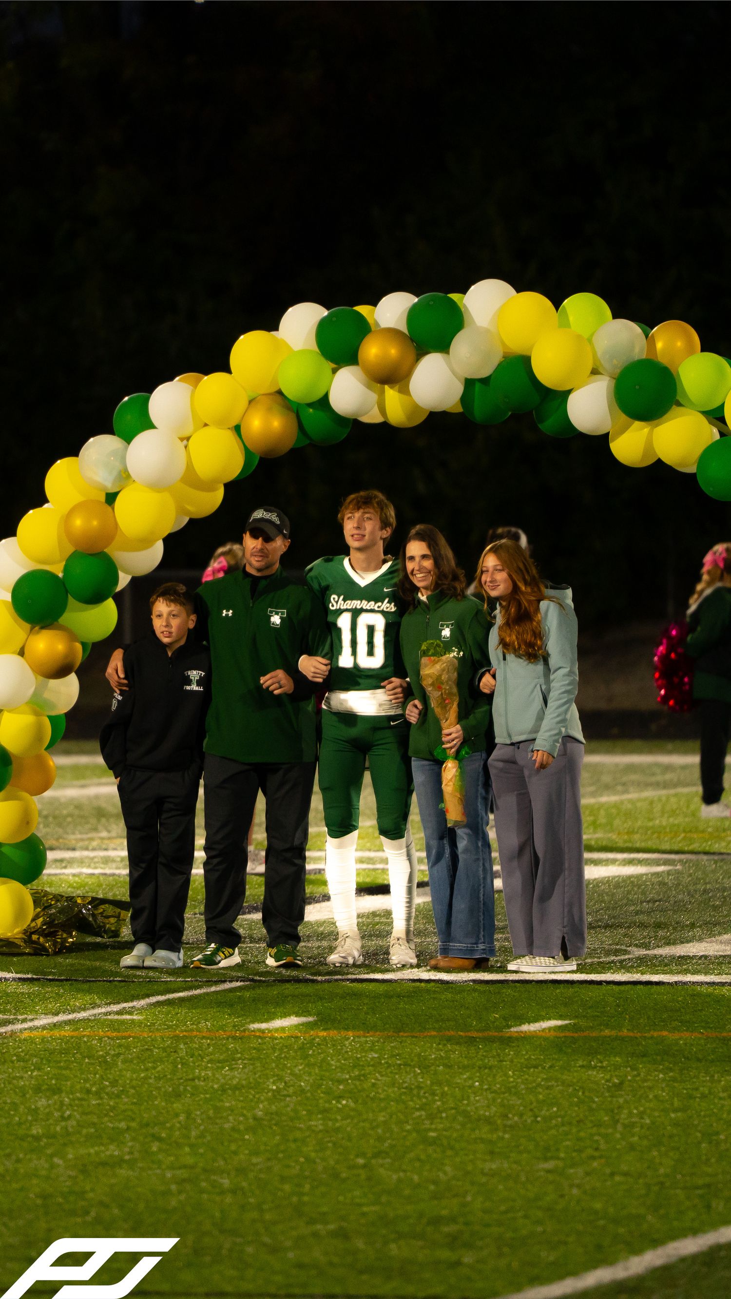 Shamrocks football player #10 poses with family under green and yellow balloon arch during senior night ceremony.