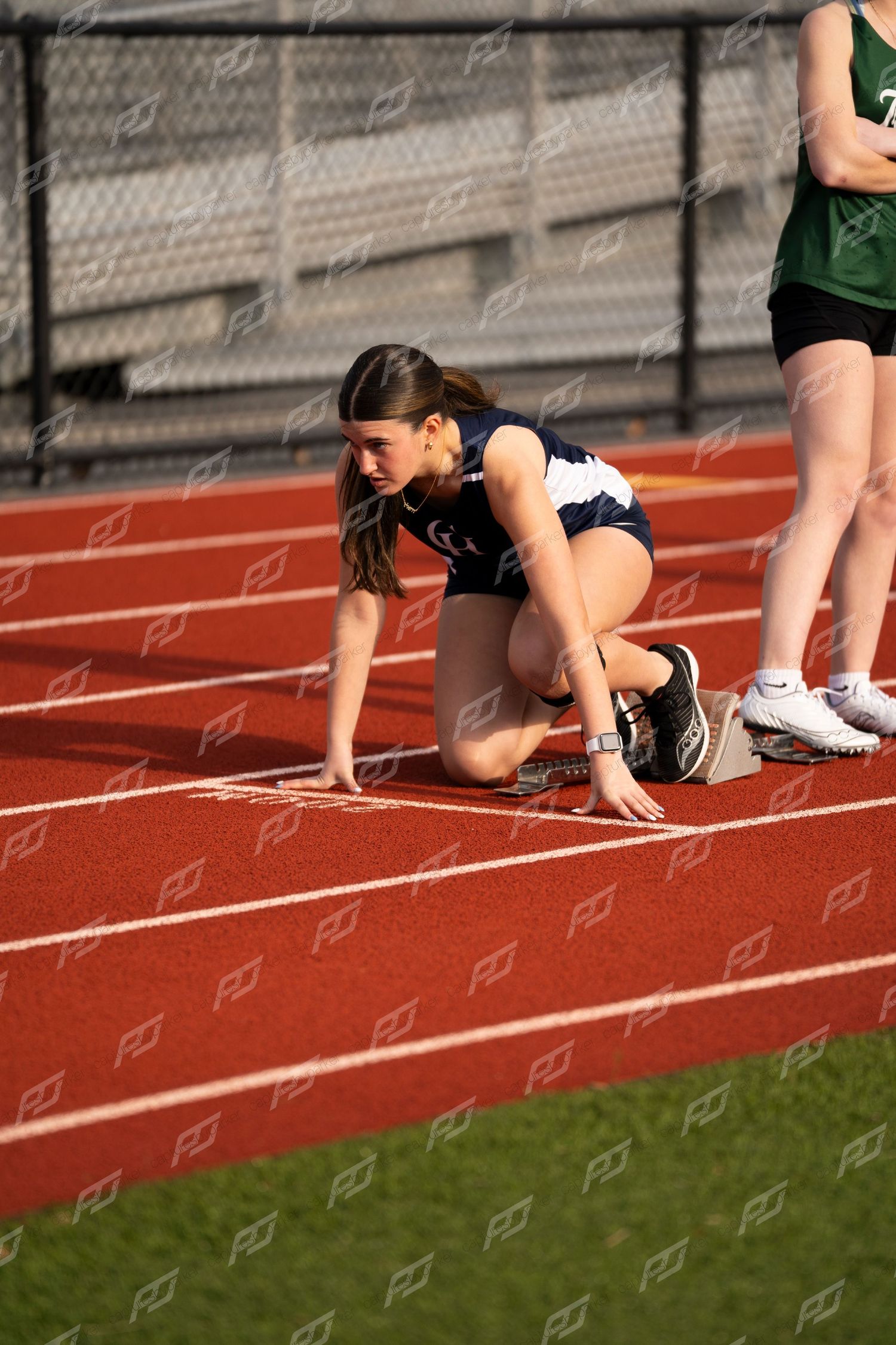 Female athlete in navy uniform in starting position on red outdoor track.