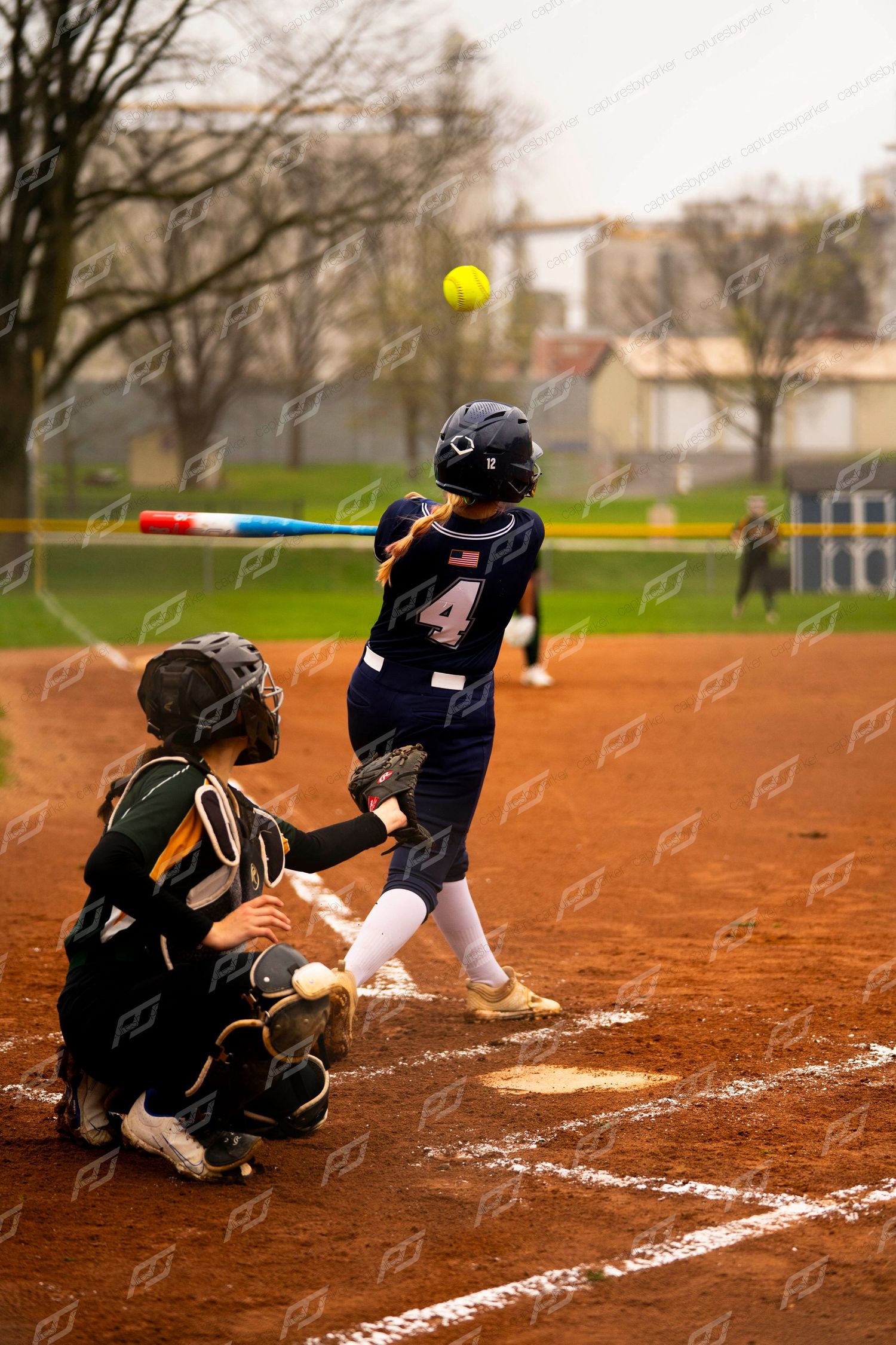 Softball batter number 4 in navy uniform swings at yellow ball as catcher crouches during a game.