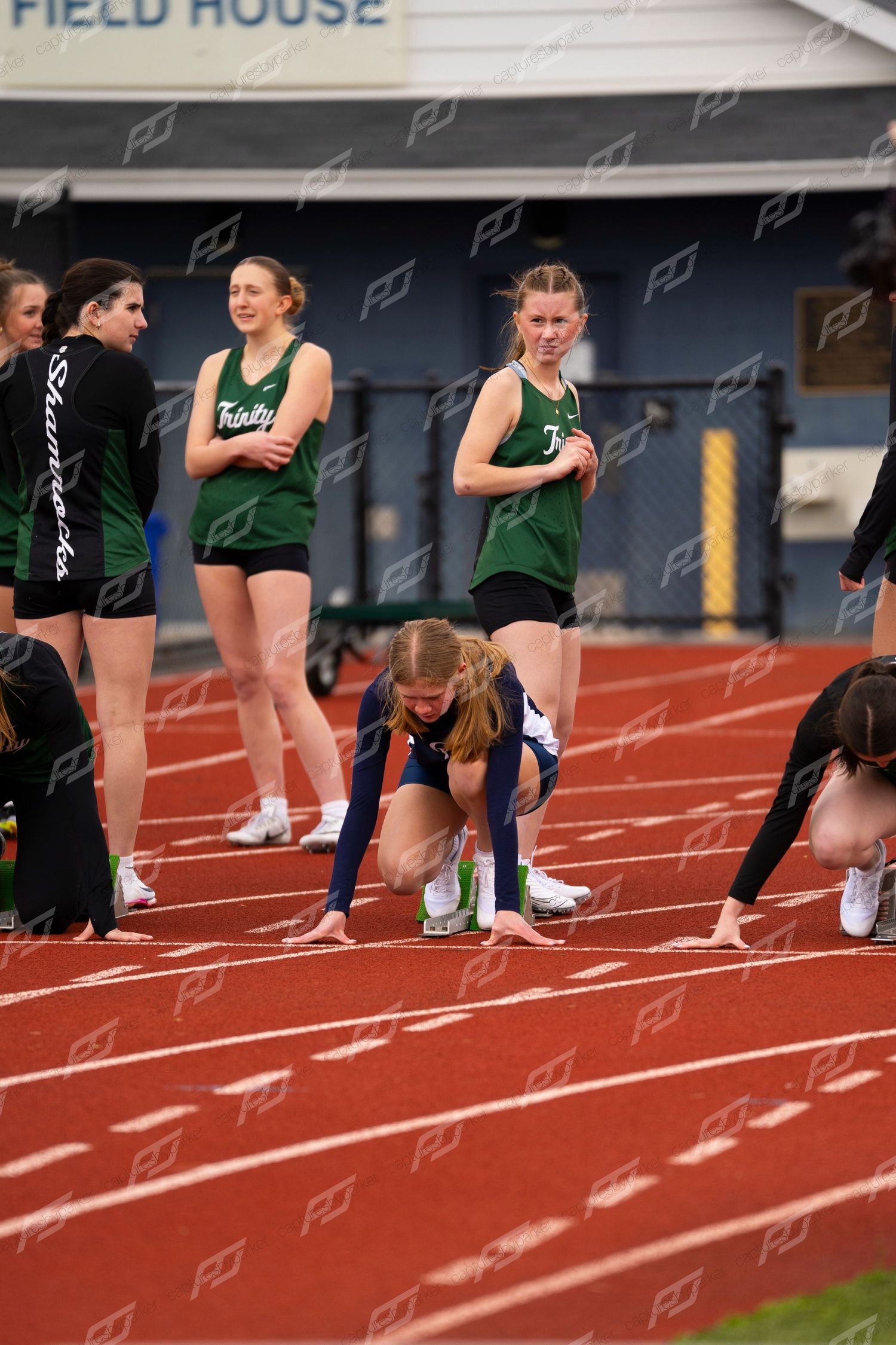 Female track athletes in green Trinity uniforms prepare at starting blocks on red outdoor track.