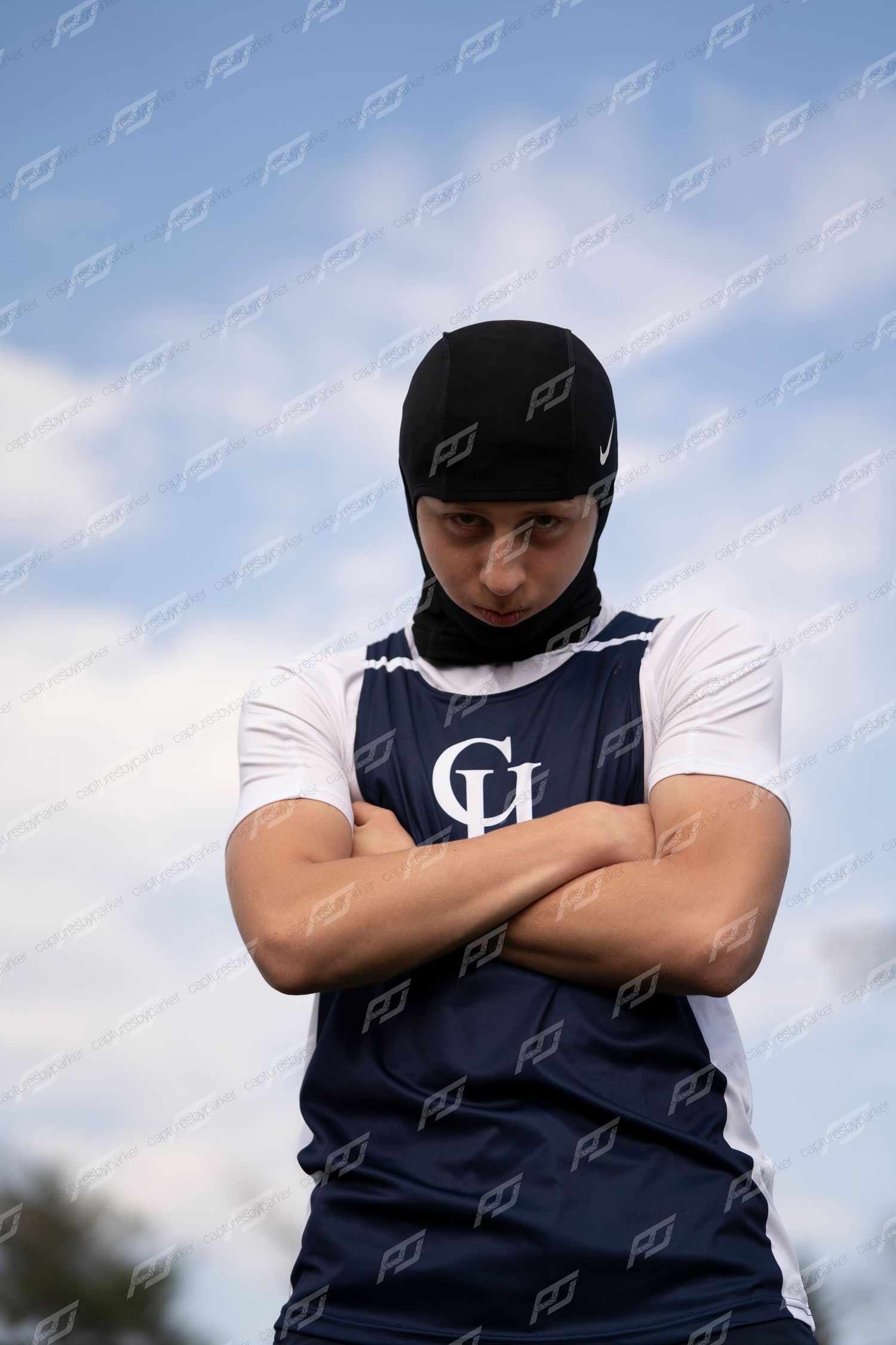 Male track athlete in navy CH vest and black head covering stands with arms crossed.