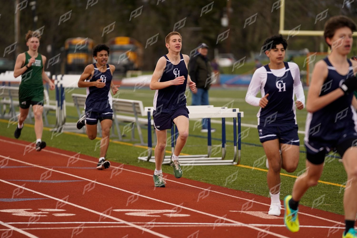 Group of male runners in navy uniforms competing in a track race with hurdles nearby.