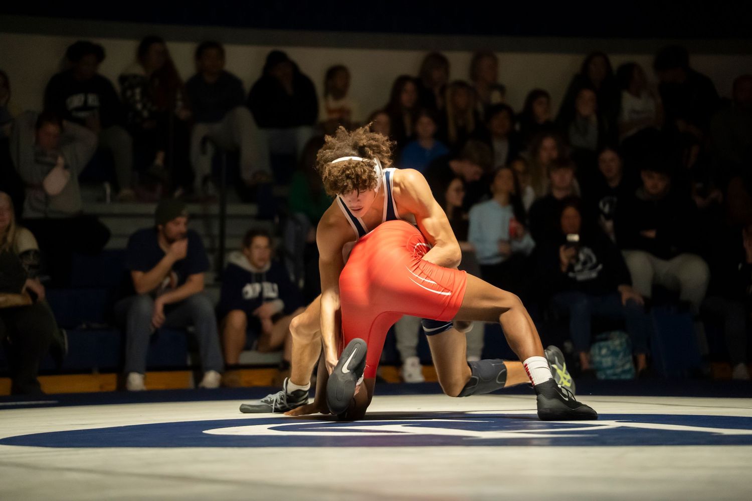 Wrestler in blue singlet lifts opponent in red during an intense wrestling match before a crowd.