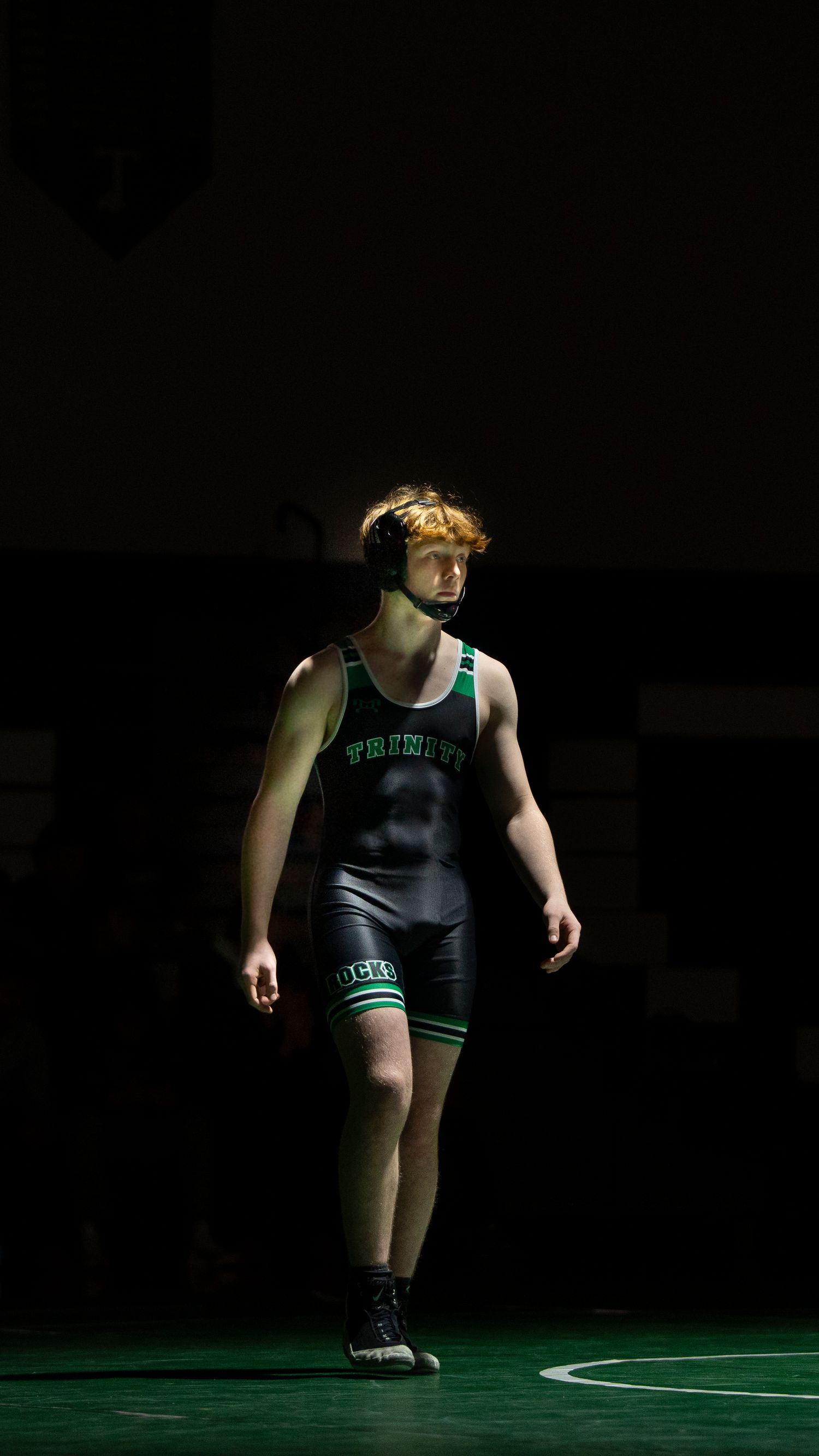 Trinity wrestler in black and green singlet walks across illuminated mat before match.