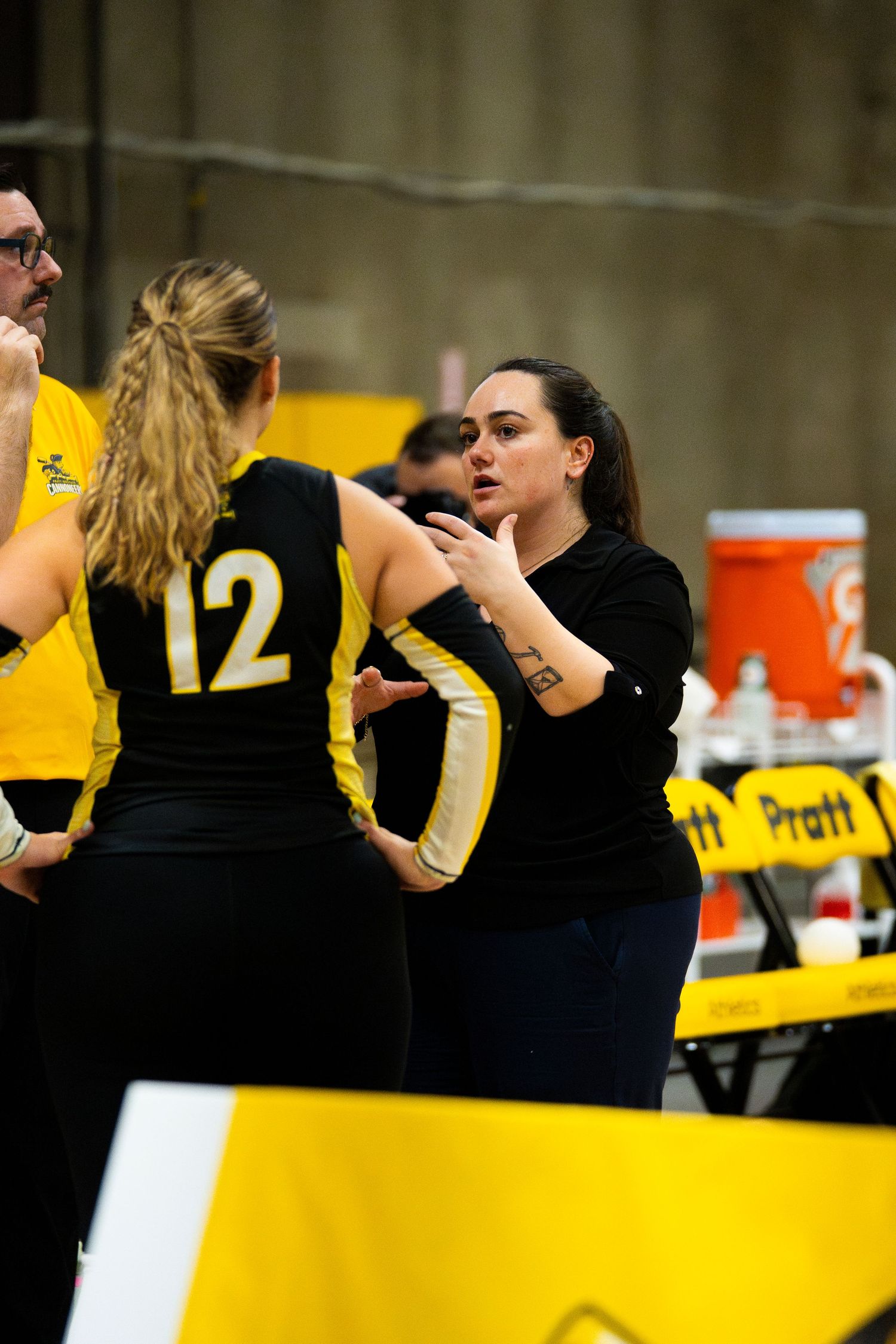 A volleyball coach in black speaks to player number 12 in a black and yellow uniform during a timeout at a Pratt sports event.