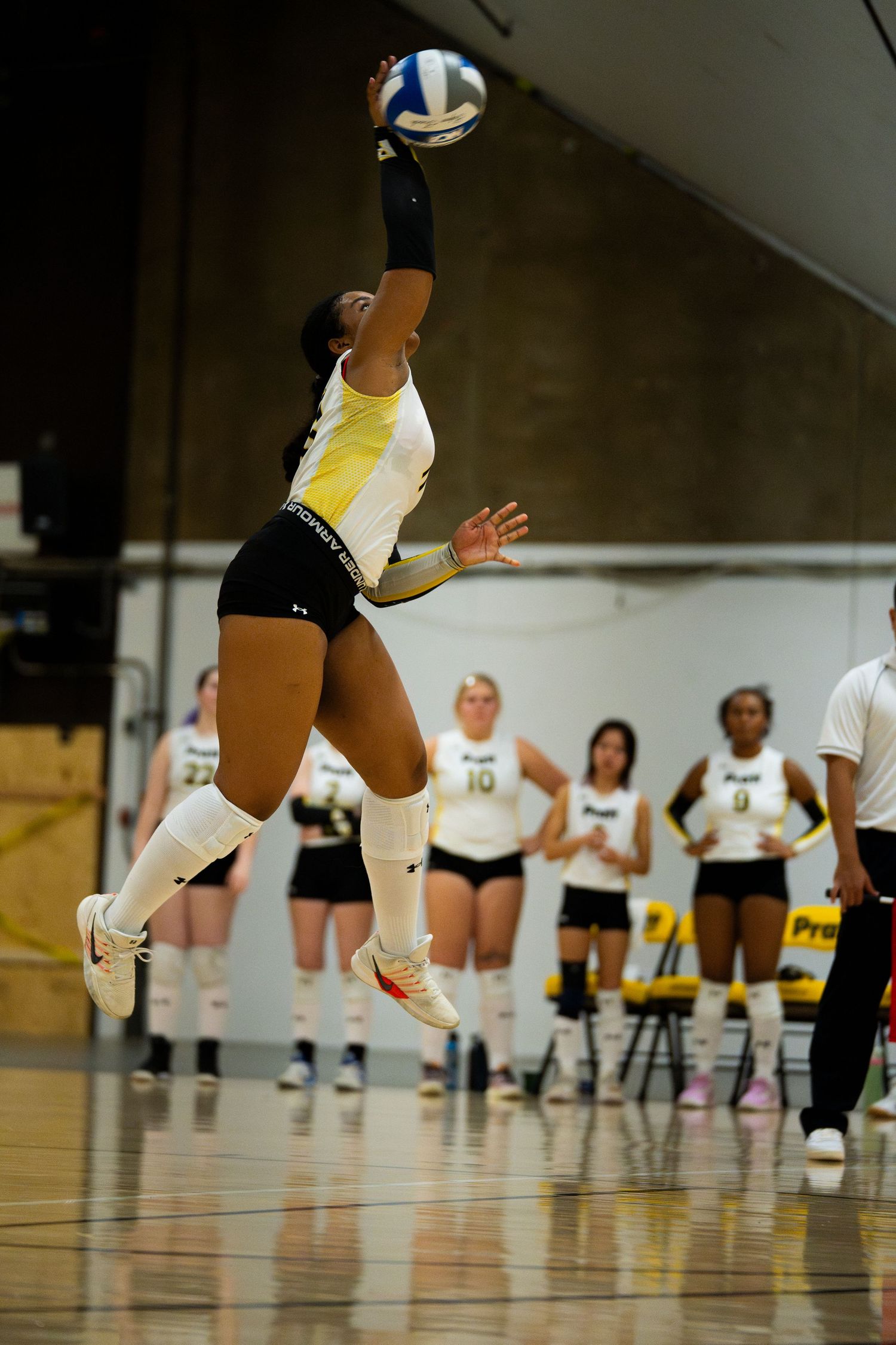 Female volleyball player leaps to serve ball during indoor match, teammates watching in background.