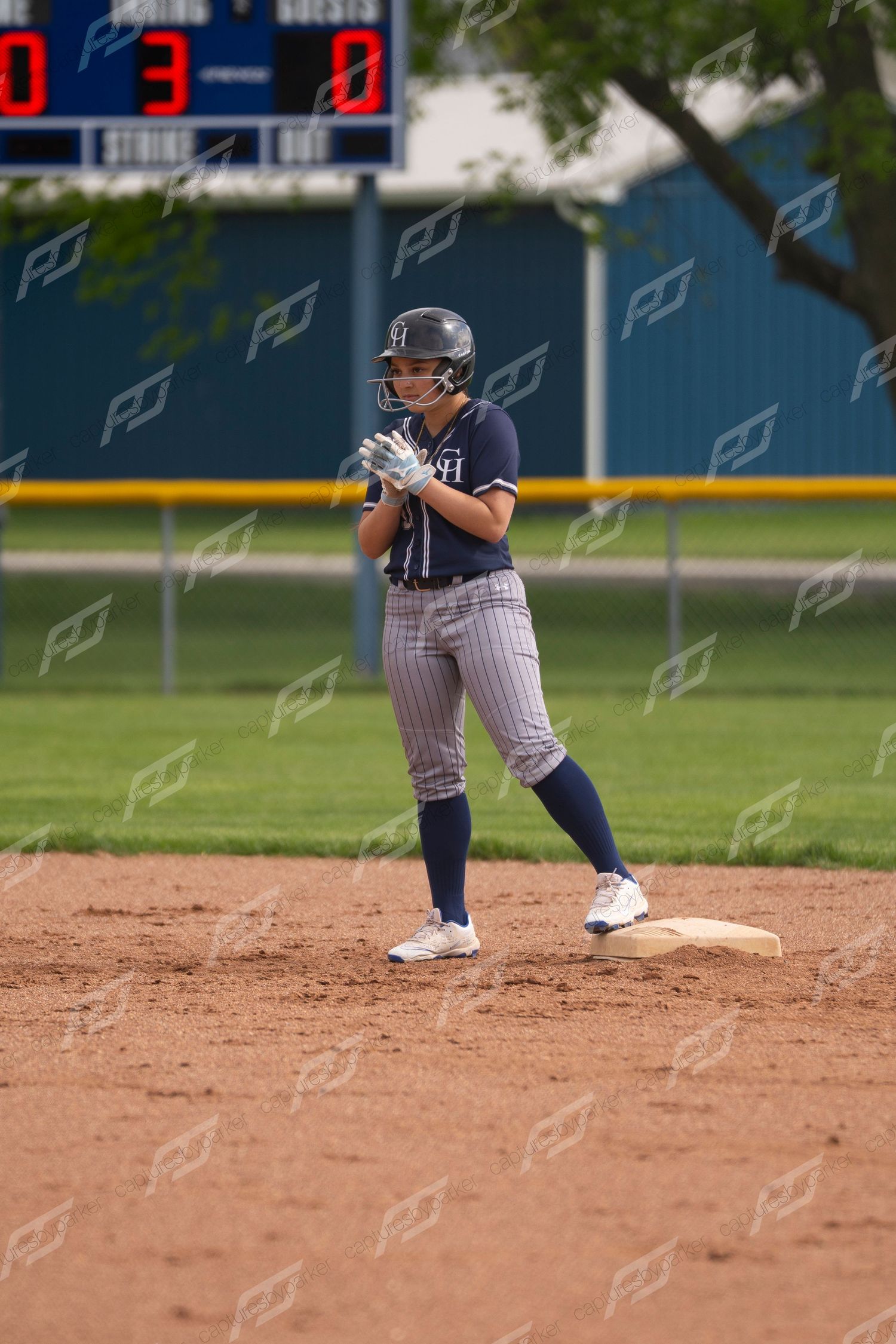 Female softball player in navy pinstripe uniform stands ready on base with scoreboard showing 3-0.