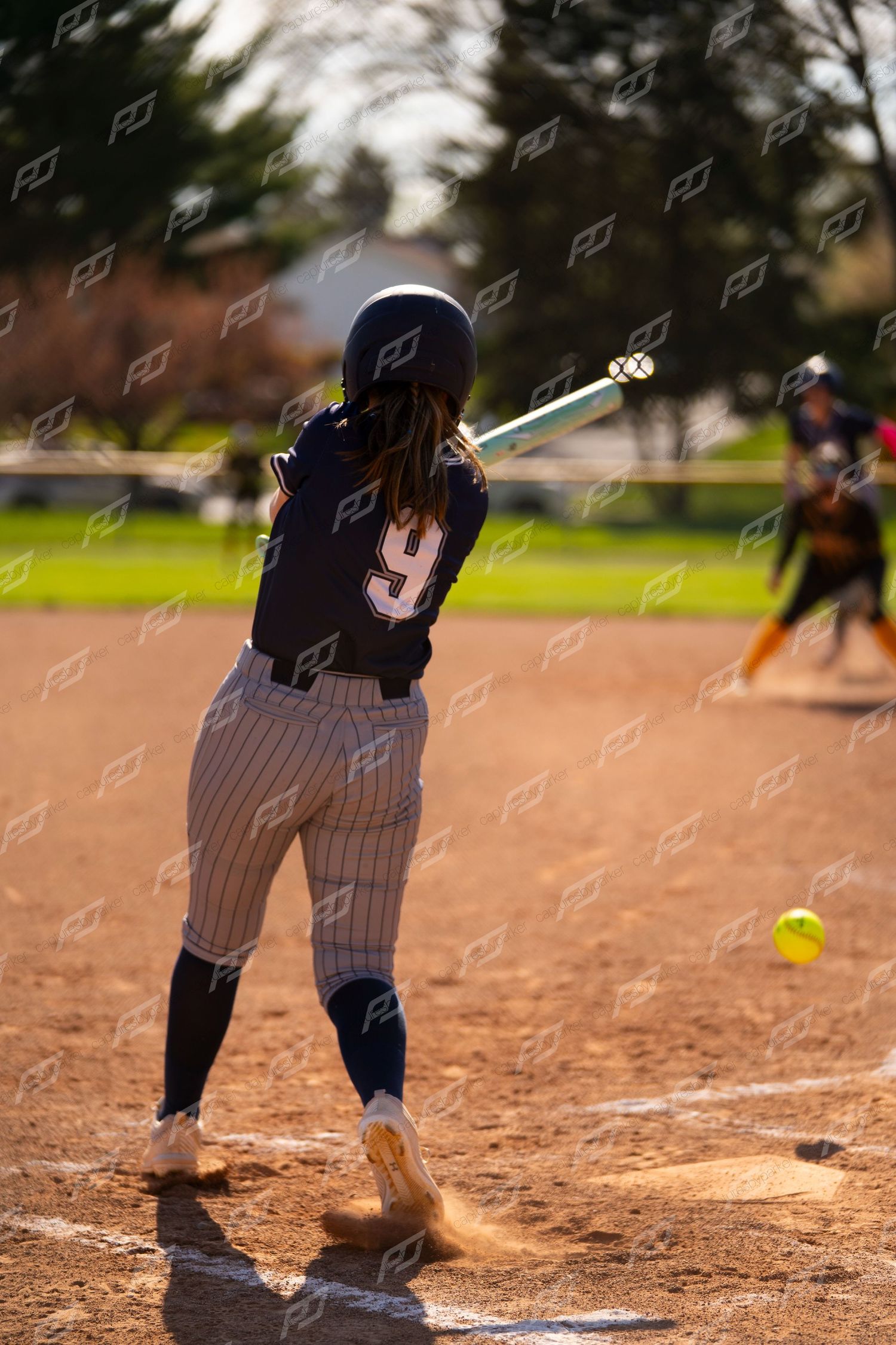 Softball player number 9 swings bat at incoming yellow ball during sunny outdoor softball game.