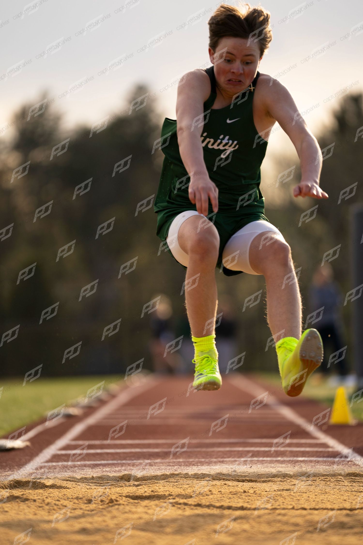 Trinity athlete in green uniform soars through air during long jump at track meet.