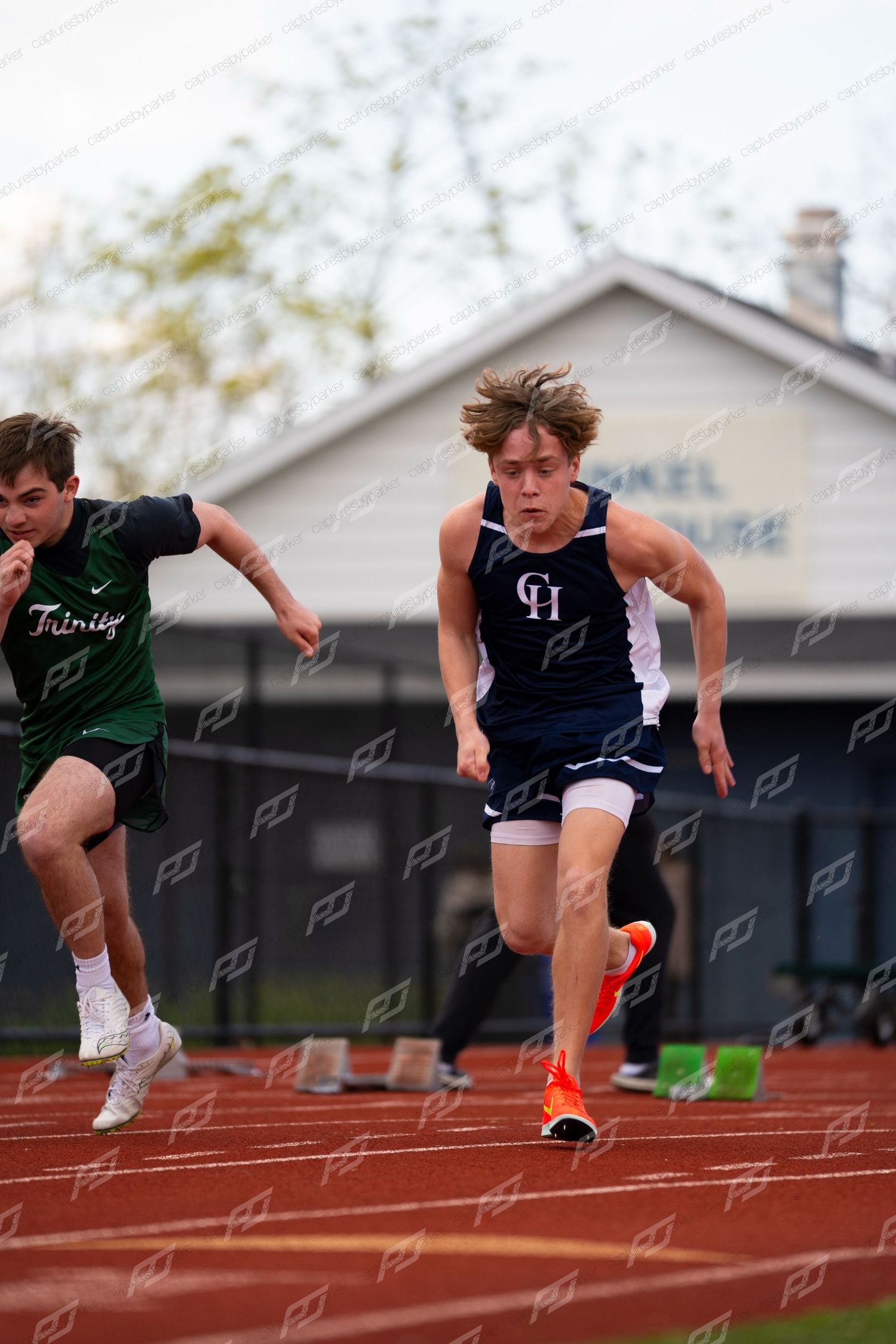 Male sprinter in GH blue uniform leads Trinity competitor off starting blocks on red track.