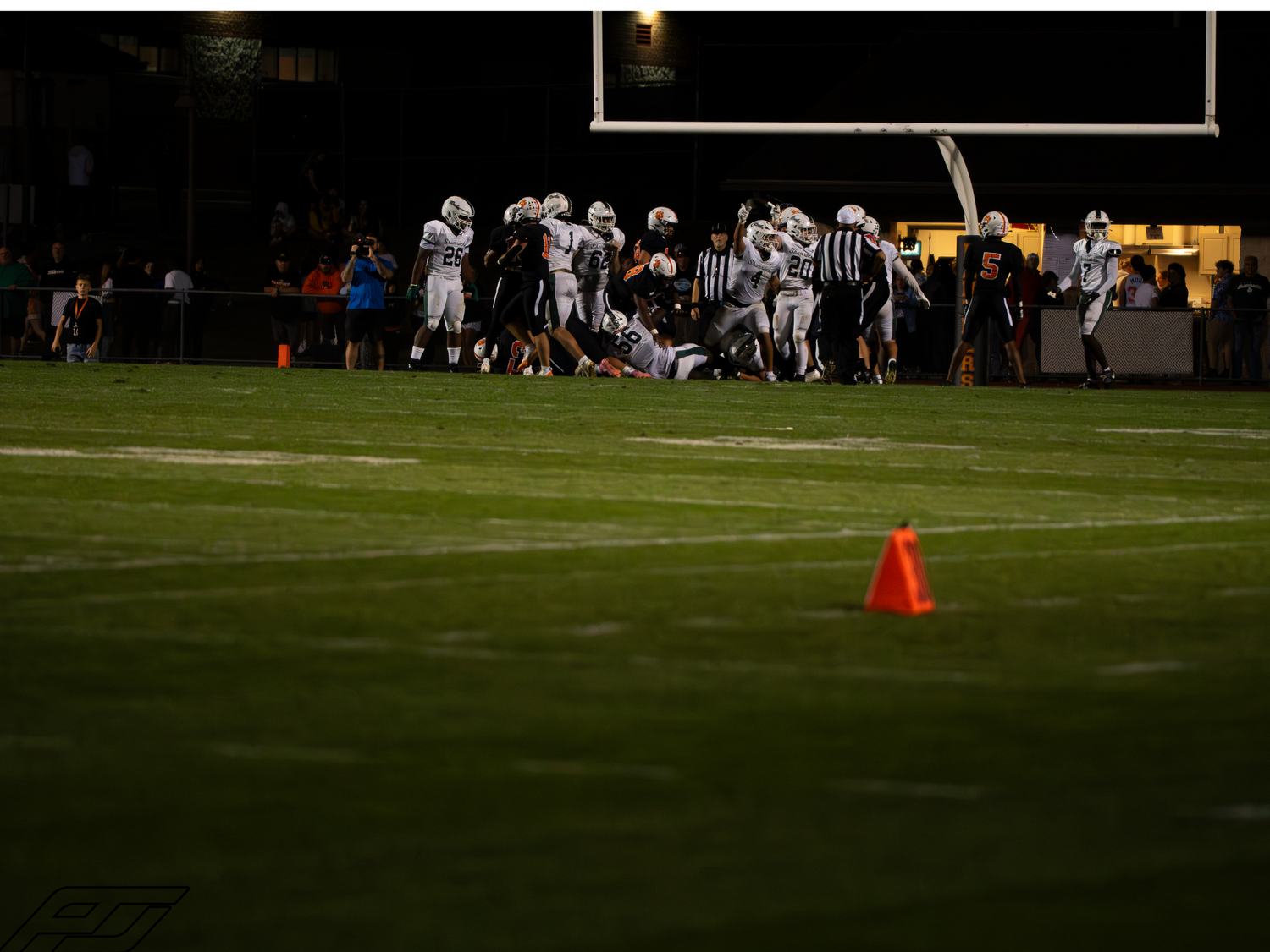 Football players pile up during a tackle on a night game field with an orange yard marker cone in the foreground.