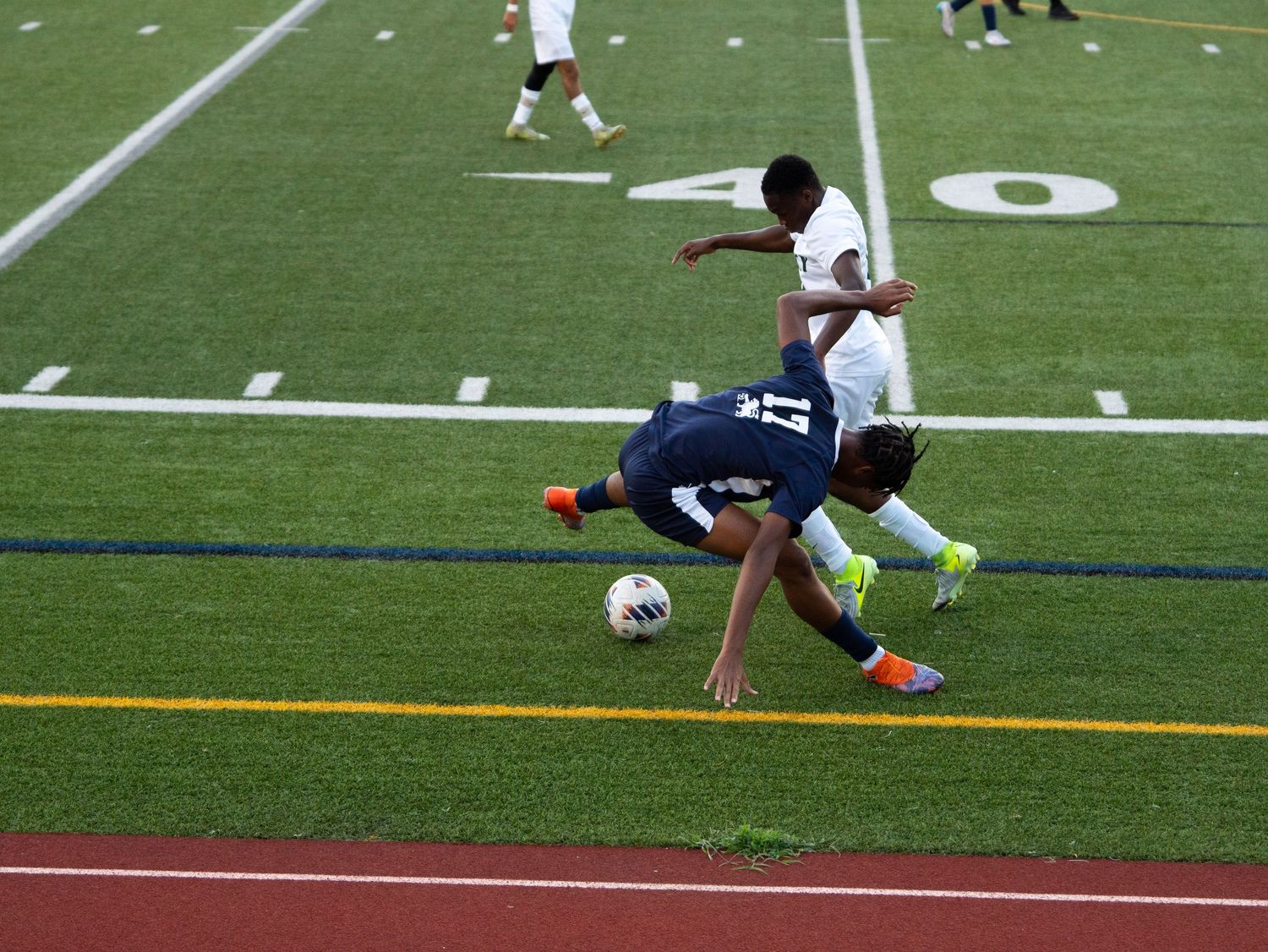 Soccer player in navy uniform falls while competing for ball against white-uniformed opponent.