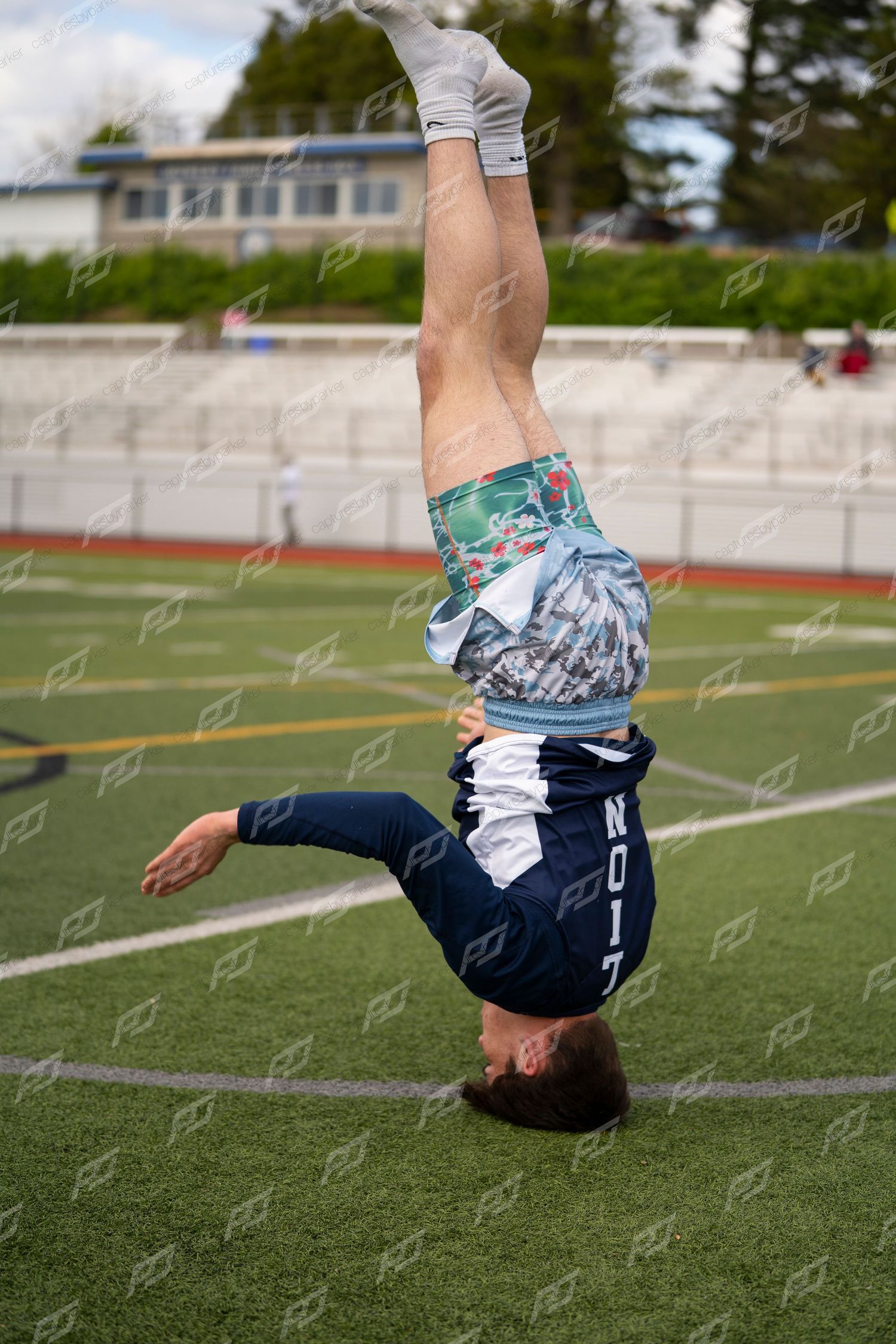 Male athlete performing a handstand on green turf field at outdoor track and field competition.