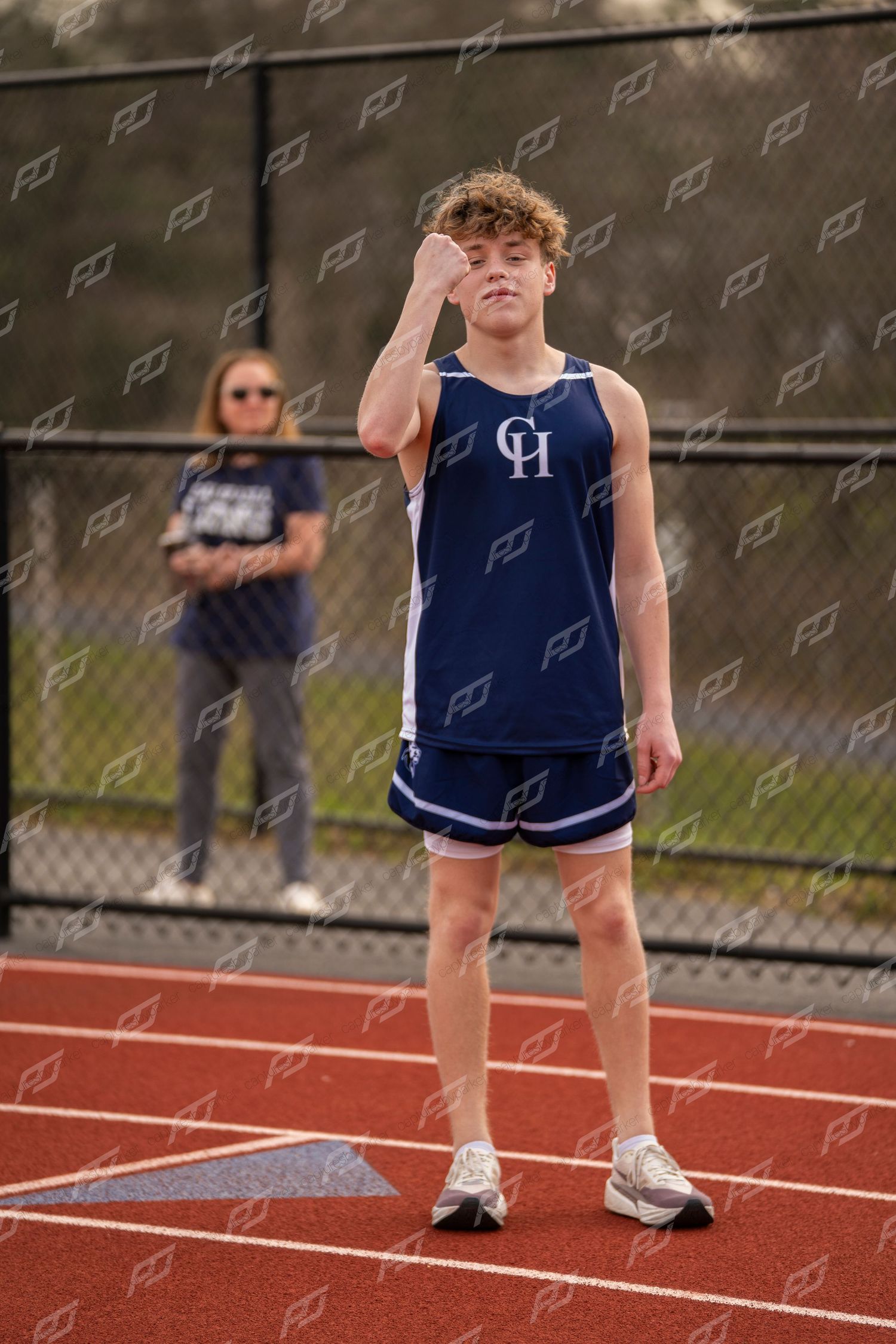 Young male track athlete in navy CH uniform stands on red track shielding his eyes.