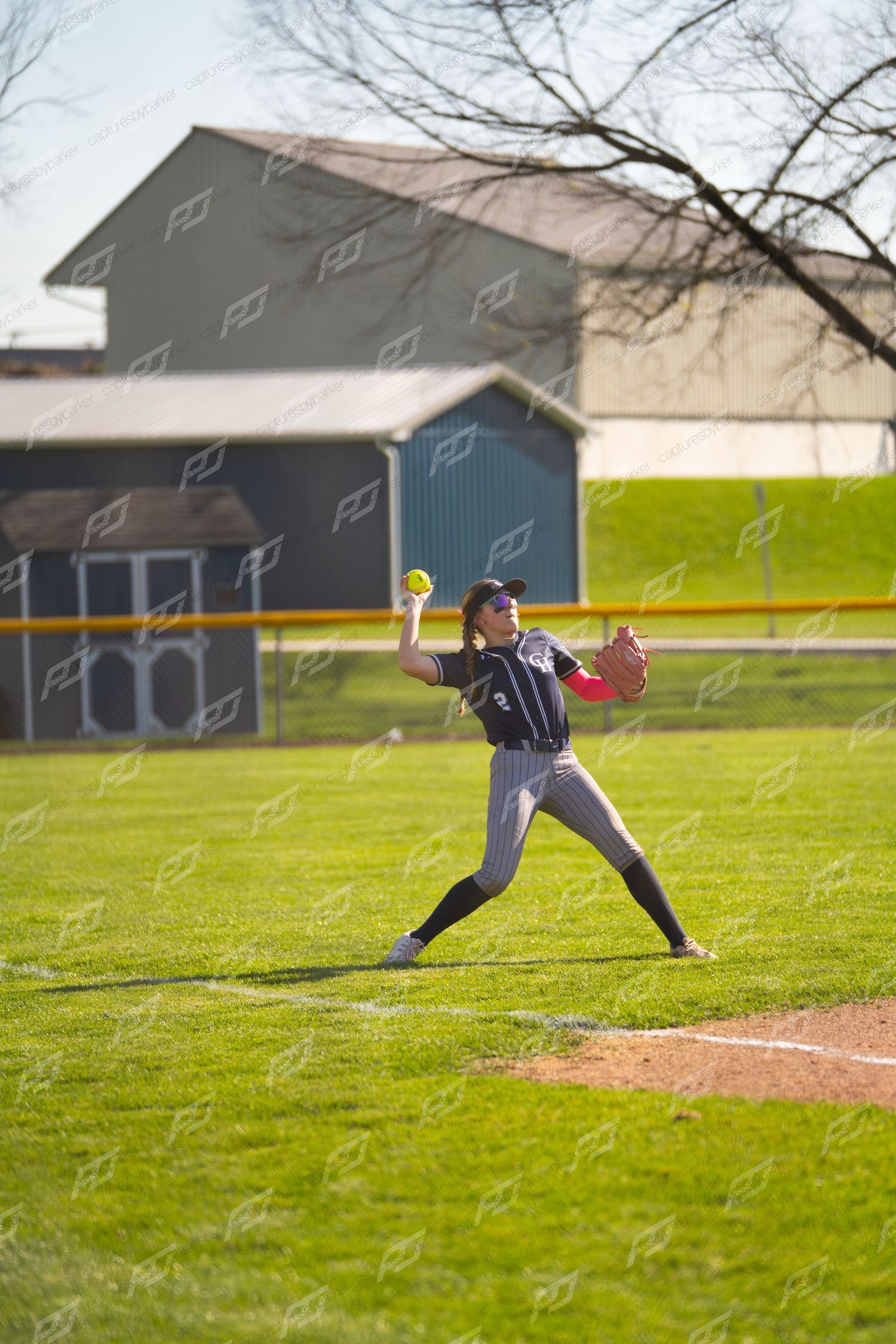 Female softball player in navy uniform winds up to throw ball during an outdoor field game.