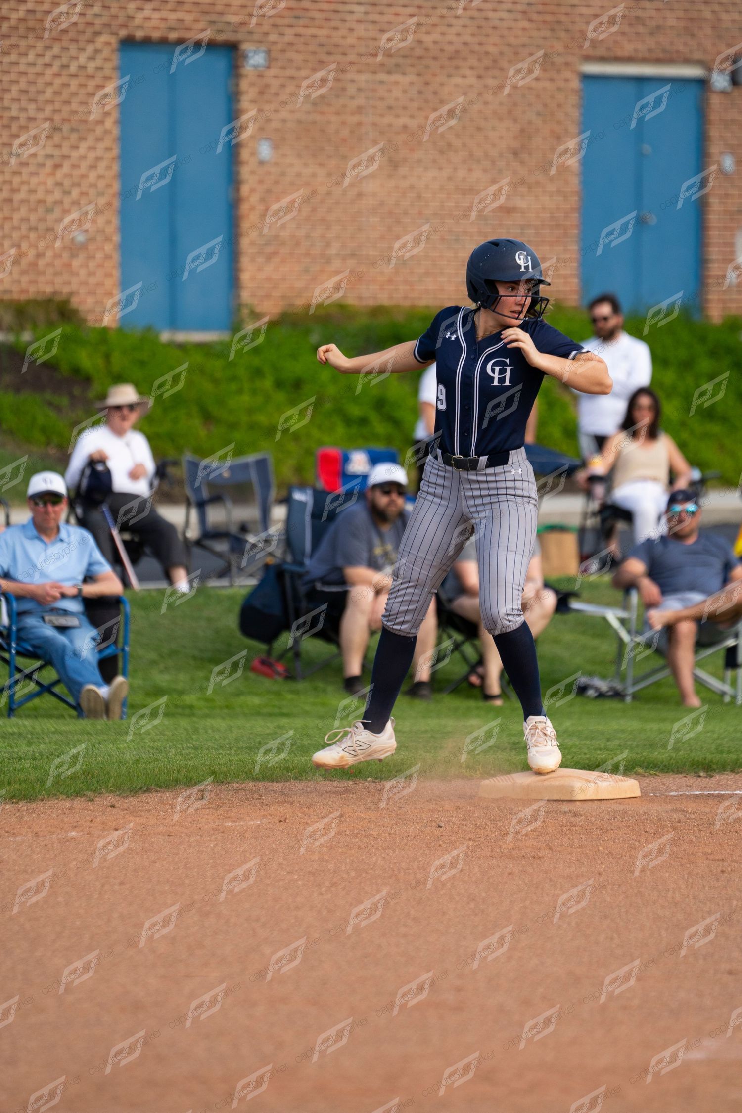 Softball player in navy pinstripe uniform steps on base during a game with spectators watching in background.
