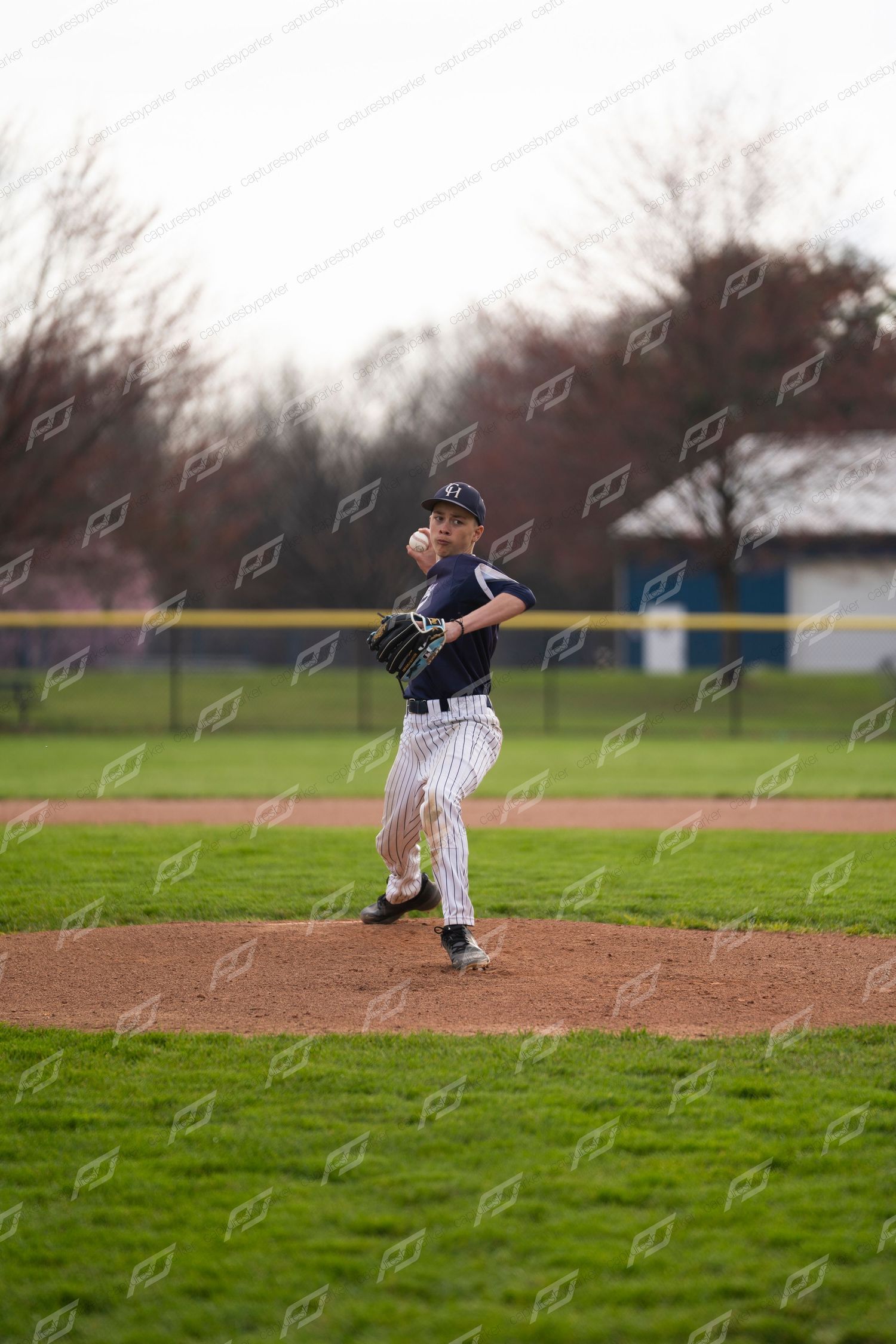 Youth baseball pitcher in navy pinstripe uniform winding up to throw from the mound on a cloudy day.