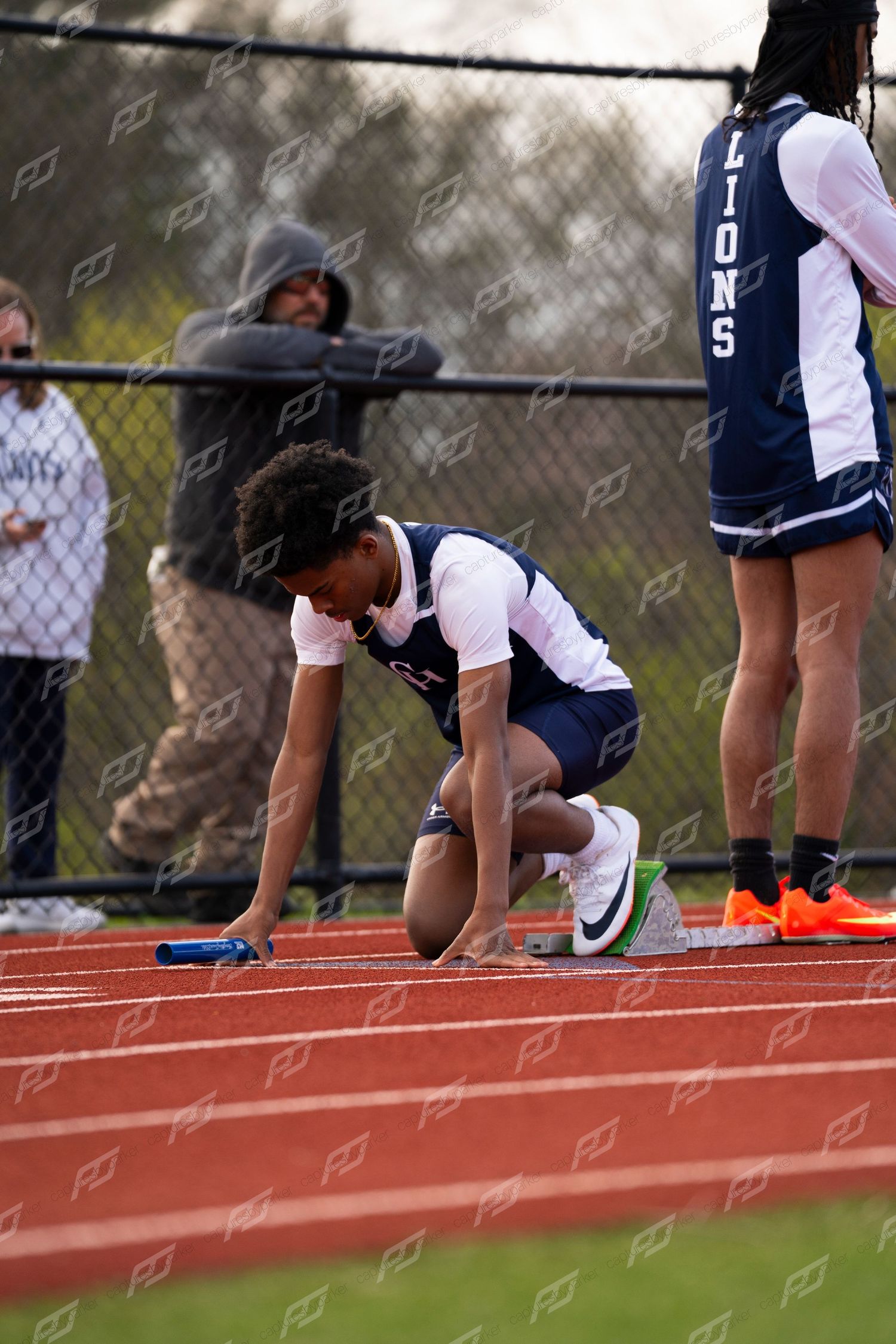 Young male relay runner crouches in starting position on red track holding blue baton ready to race.